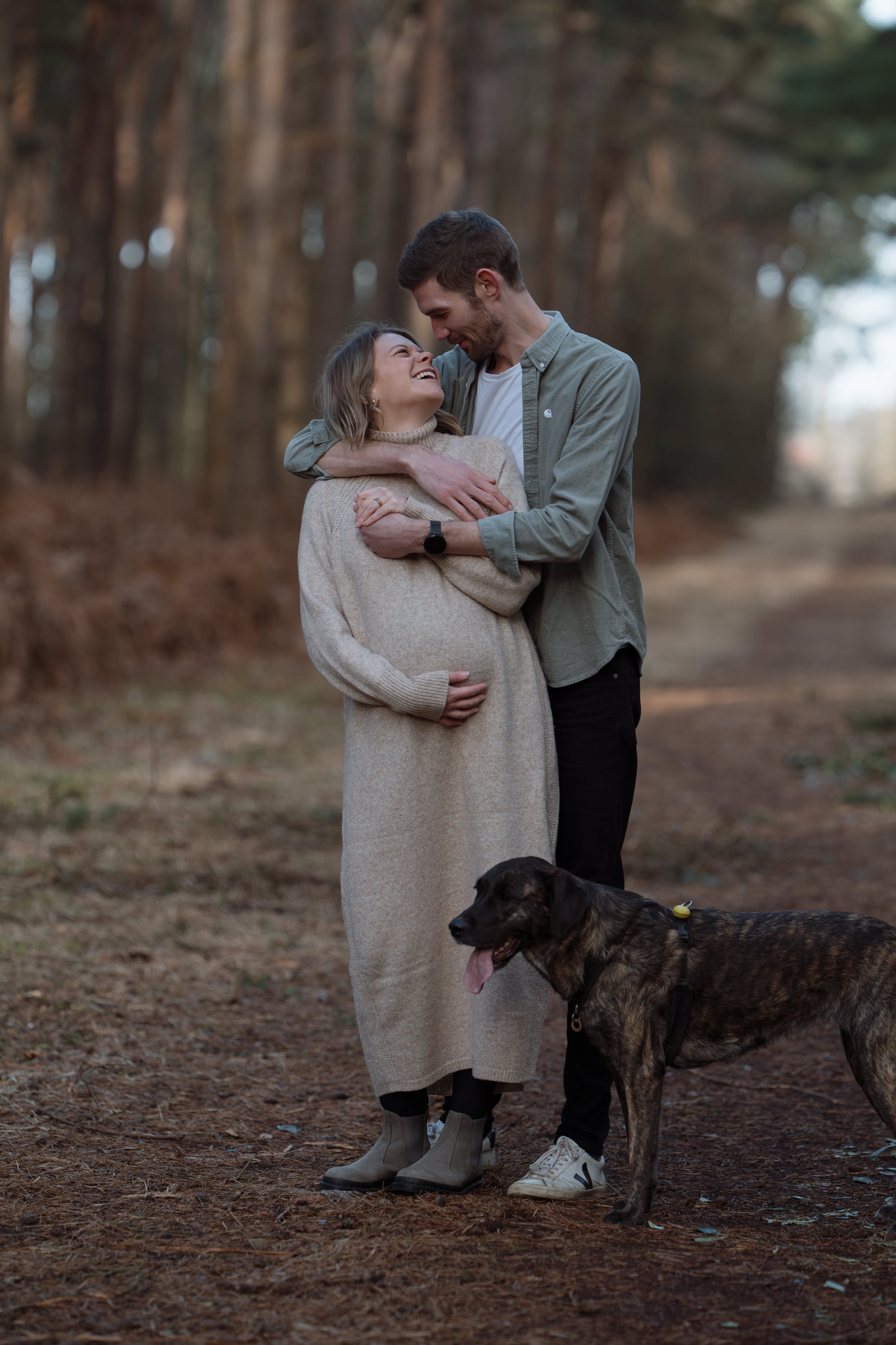 A couple standing on a forest path, embracing and smiling at each other, with a dog beside them.