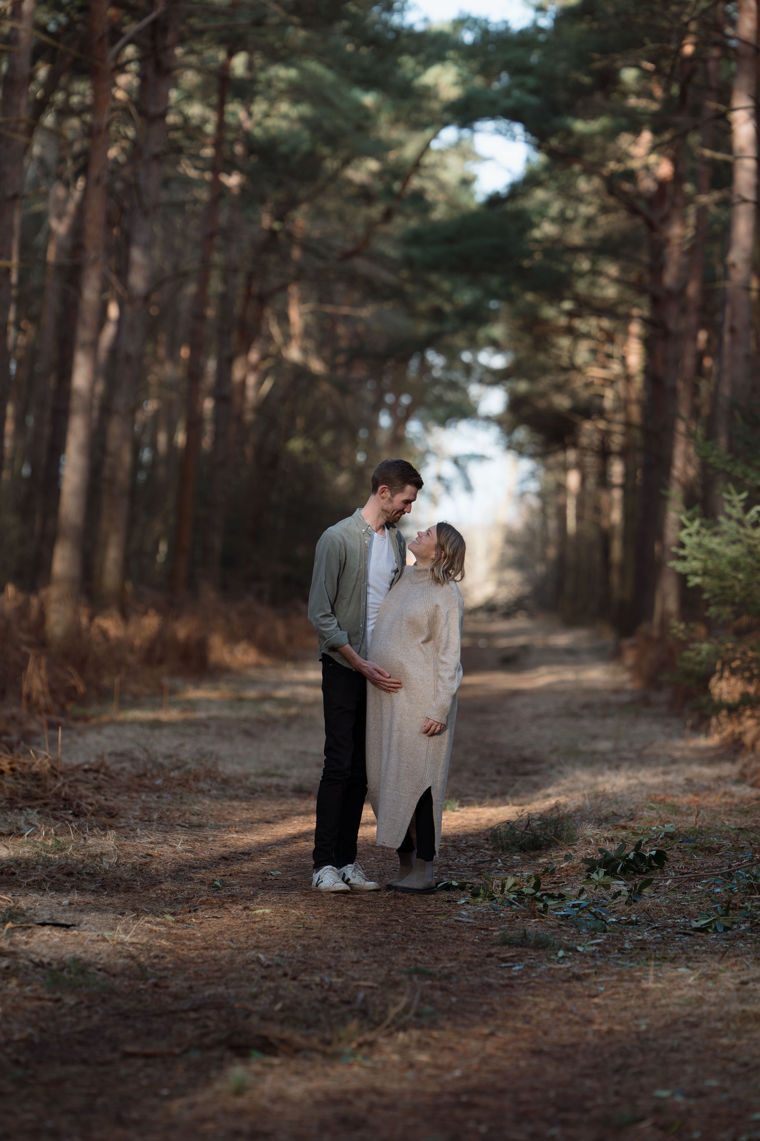 A man and woman standing close together in a forest, looking into each other's eyes and smiling, with trees in the background.