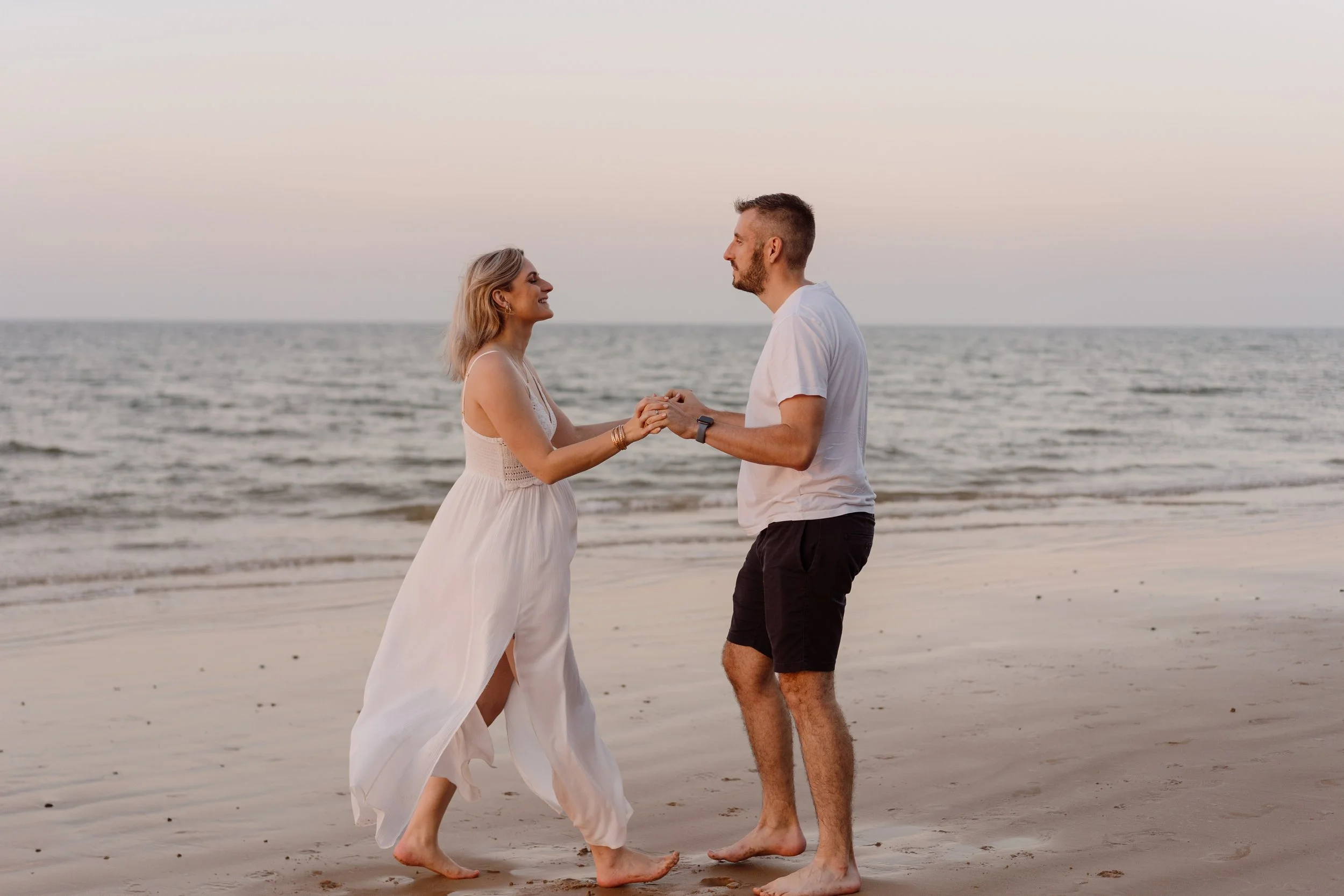 A couple holding hands on a beach at sunset, facing each other and smiling.