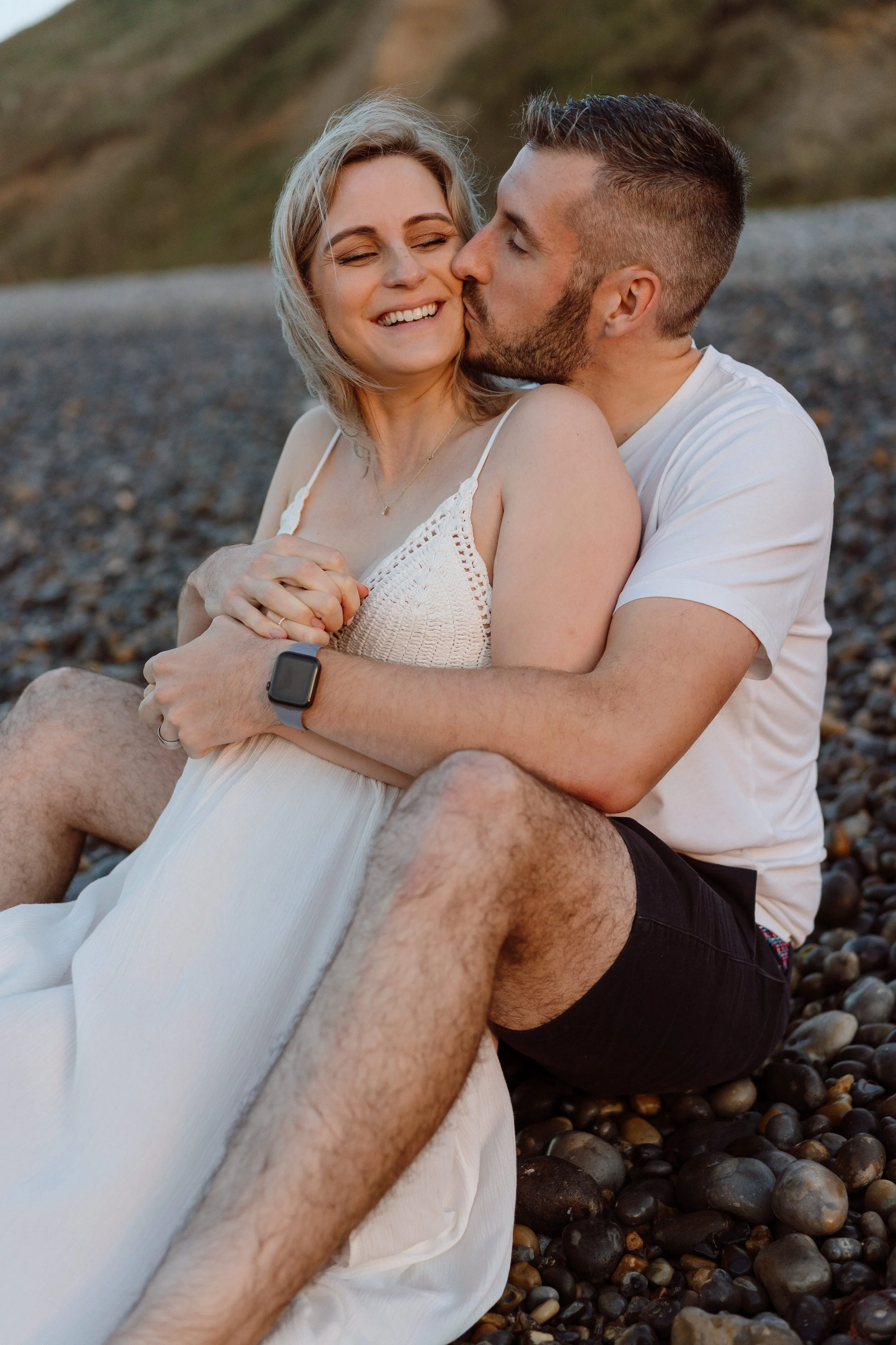A young couple sitting on a pebble beach, embracing each other, with the man kissing the woman on the cheek. The woman is smiling and holding hands with the man. They are dressed casually, with the woman in a white dress and the man in a white t-shir