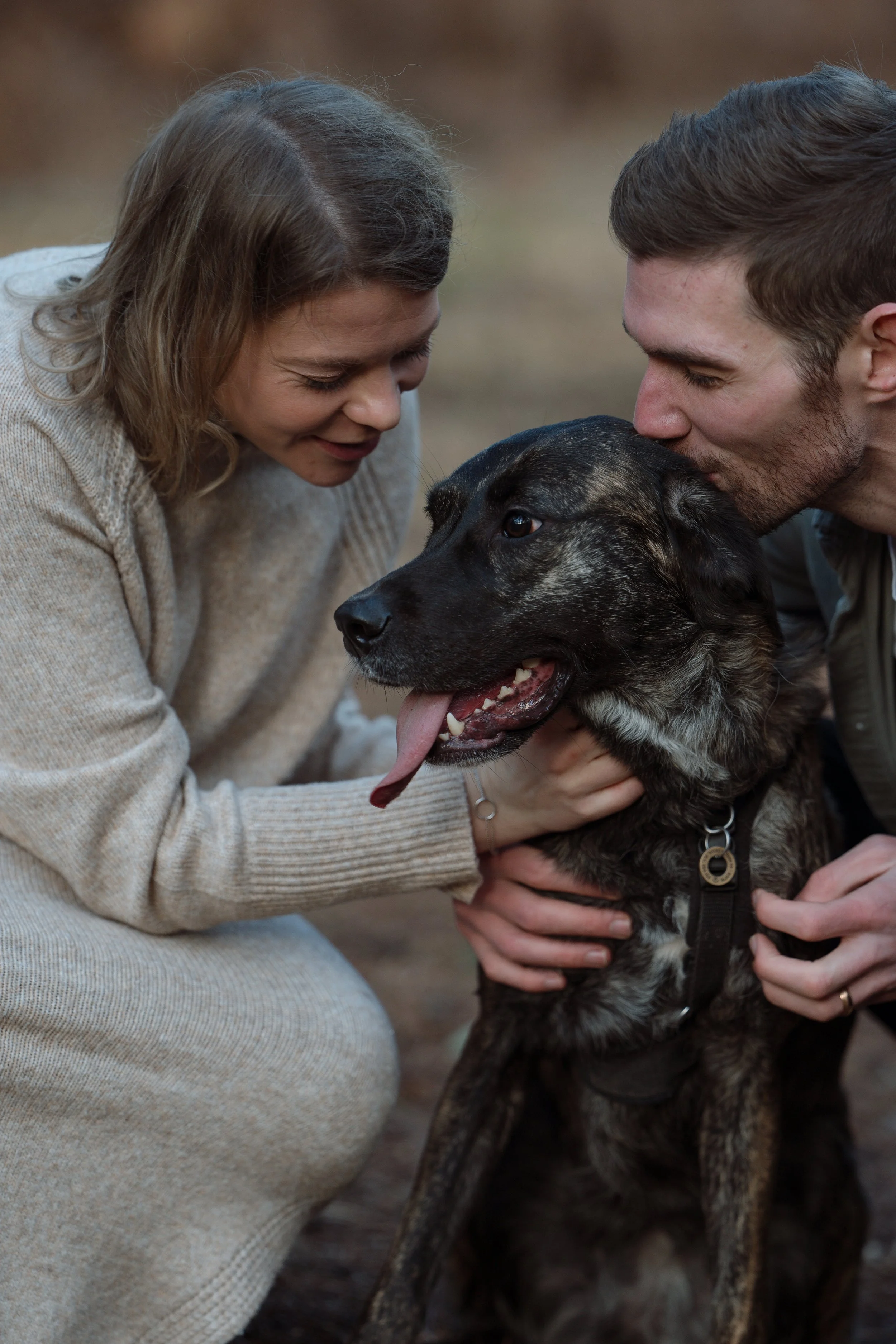 A woman and man affectionately interacting with a large black and brown dog outdoors on a dirt ground, with both people smiling and the woman holding the dog's collar.