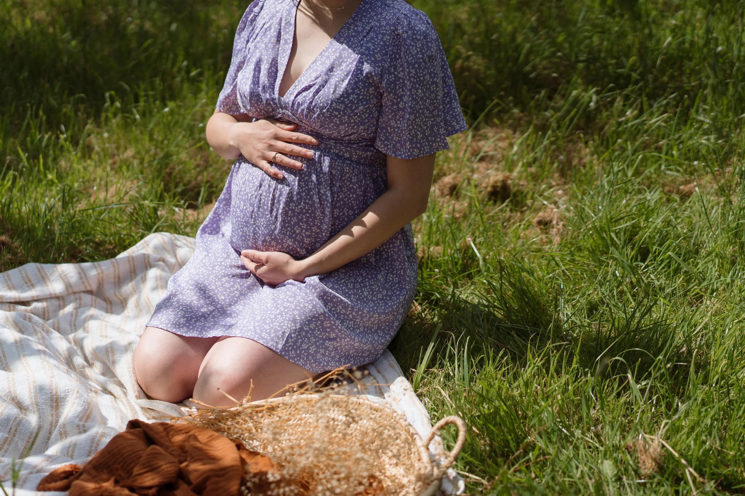 A pregnant woman wearing a purple dress with white floral pattern sitting on a blanket in a grassy outdoor area, resting her hand on her belly.