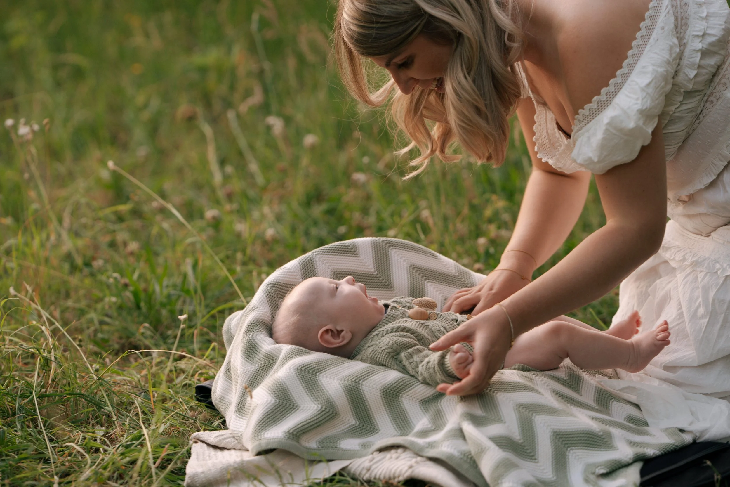 A woman in a white dress gently touching a baby lying on a blanket outdoors in a grassy field during daytime.