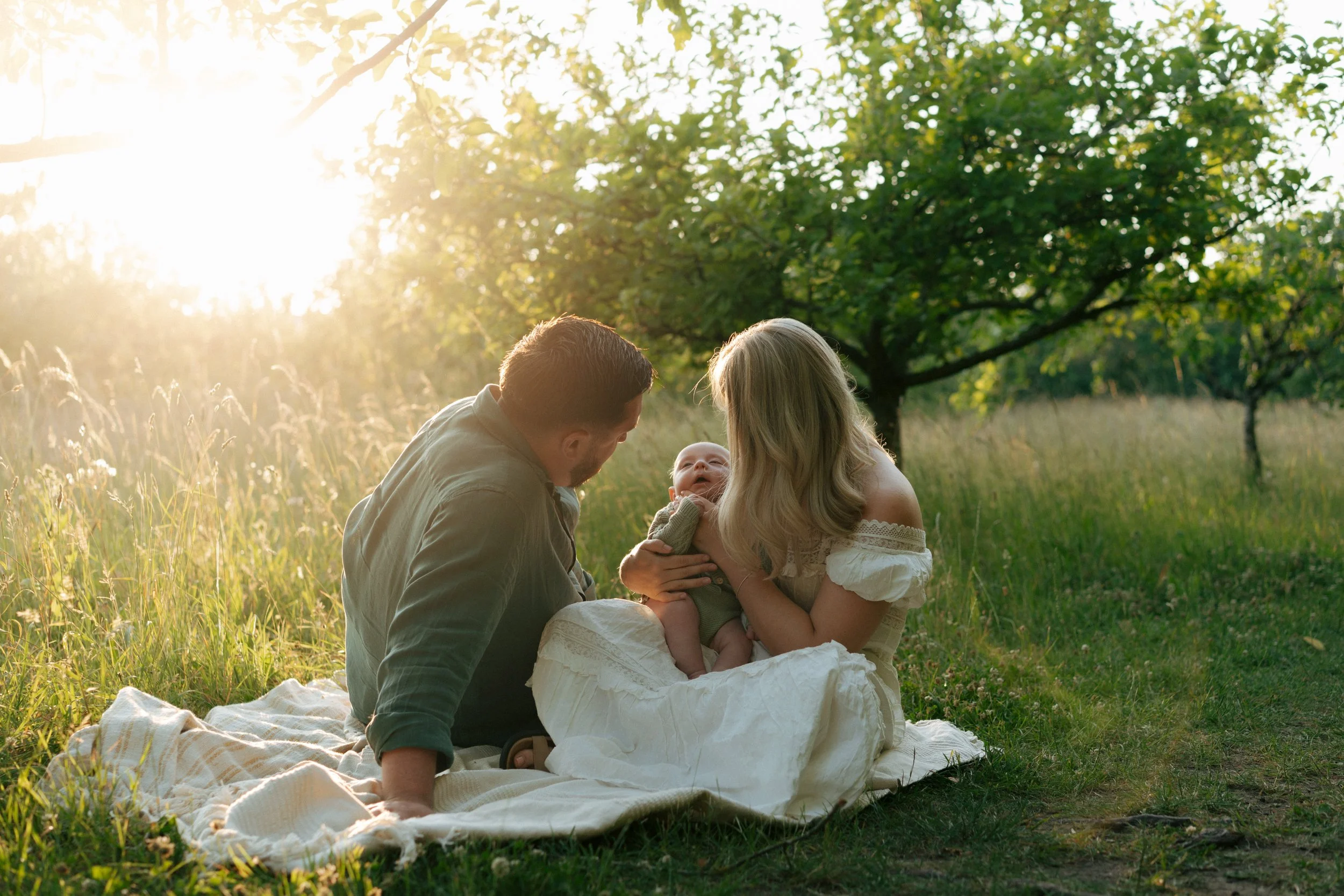 Family of three sitting on a blanket in a grassy field during sunset, with trees and sunlight in the background. The woman is holding a baby, and the man is sitting next to them.