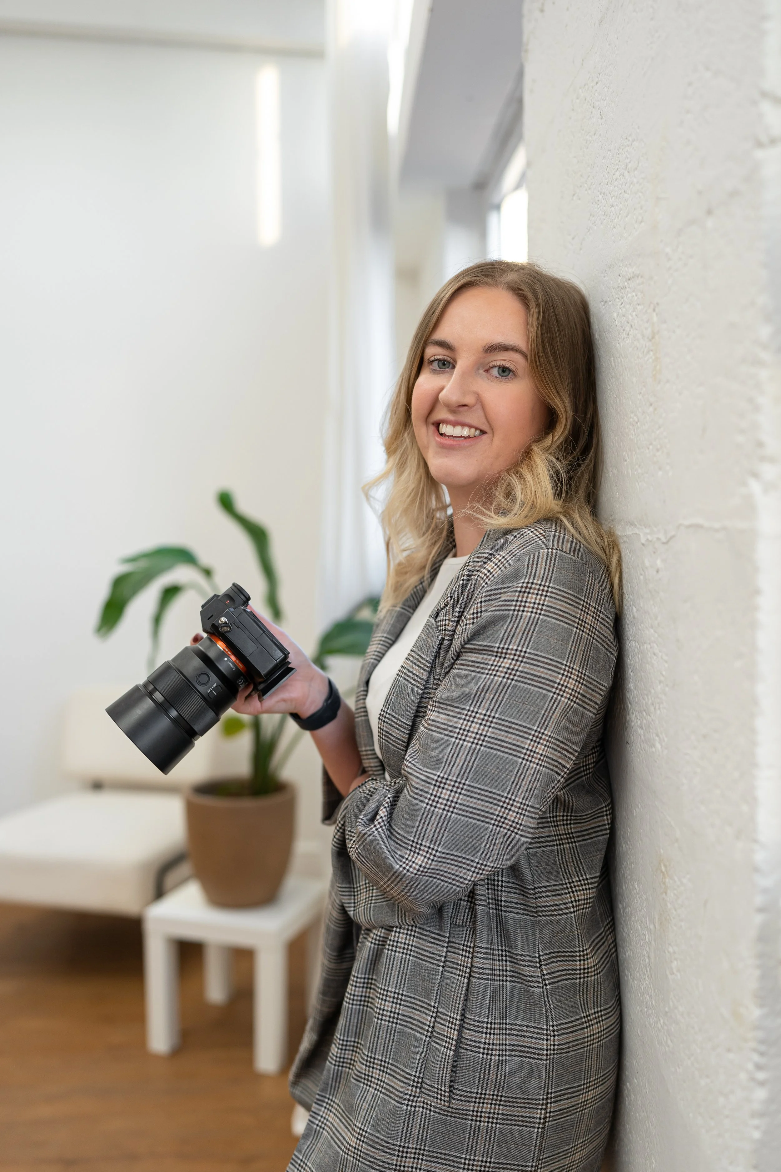 A woman with blonde, wavy hair smiling while leaning against a white wall, holding a professional camera. She is wearing a plaid blazer inside a bright, modern room with a potted plant and a white chair visible in the background.