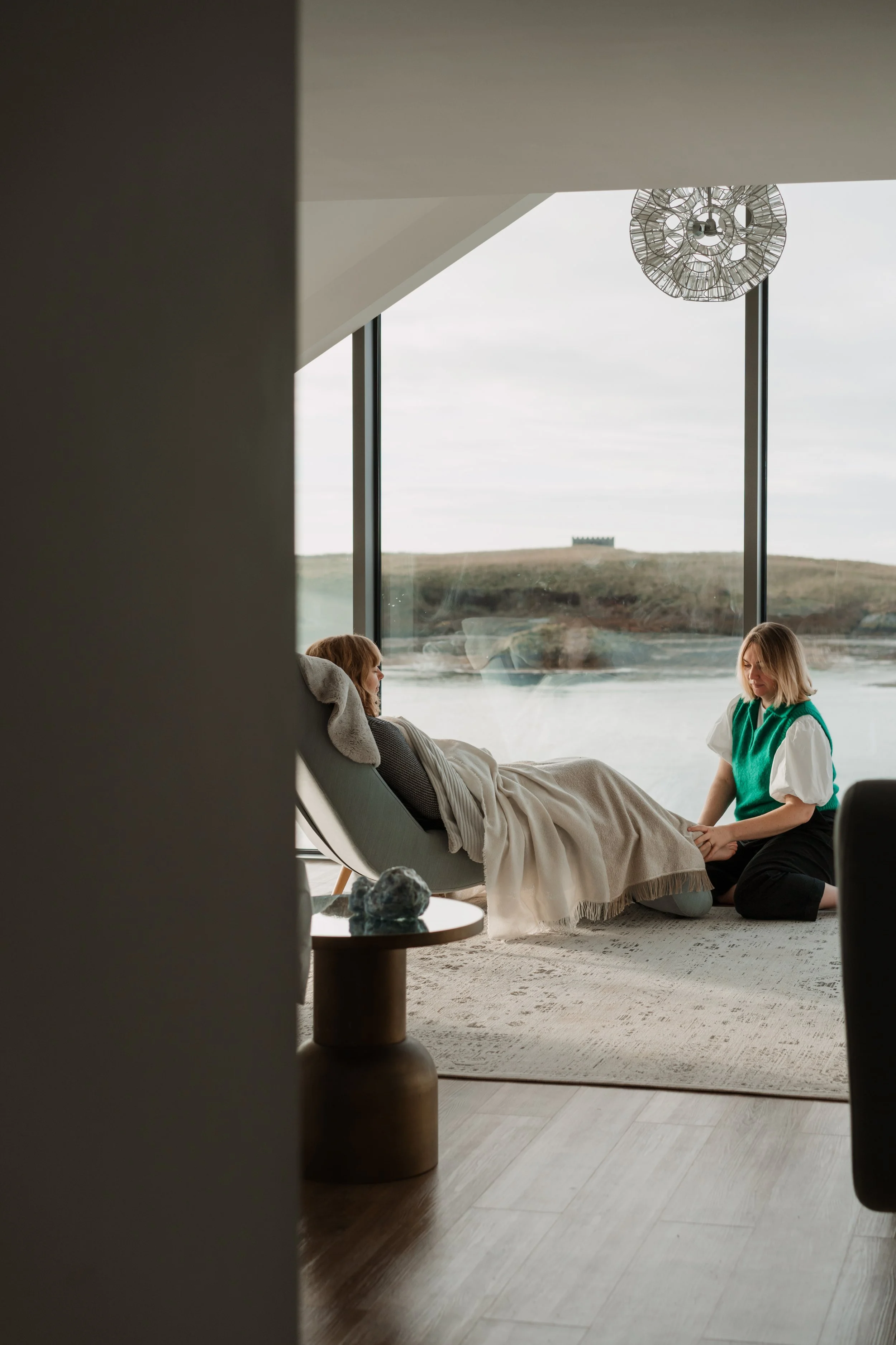 A woman sitting beside a patient reclining in a lounge chair in a modern room with large windows overlooking water and a hilly landscape, with a decorative ceiling light.