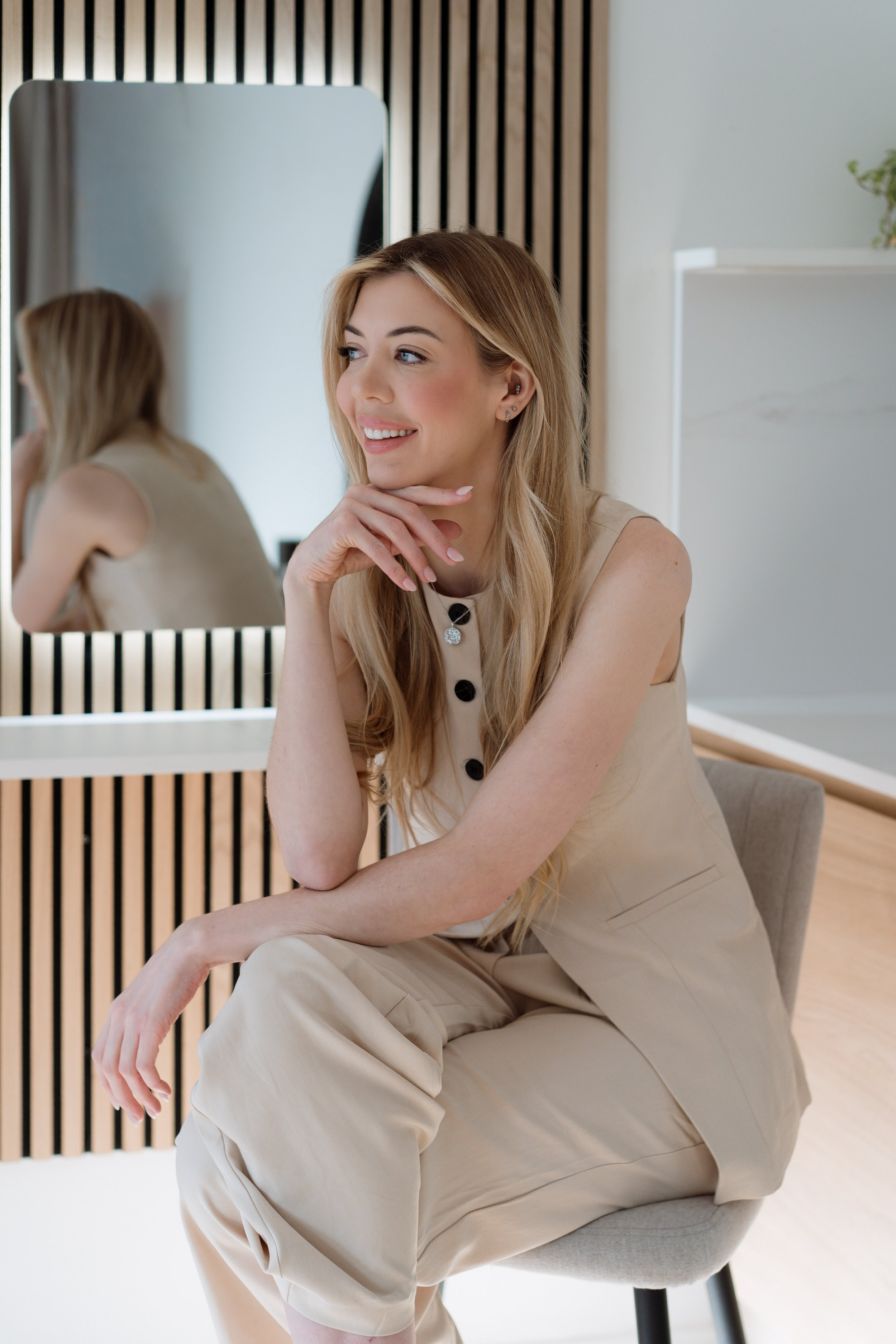 A woman sitting on a couch, smiling and looking at her phone, in a cozy living room with a framed picture, potted plant, and books in the background.