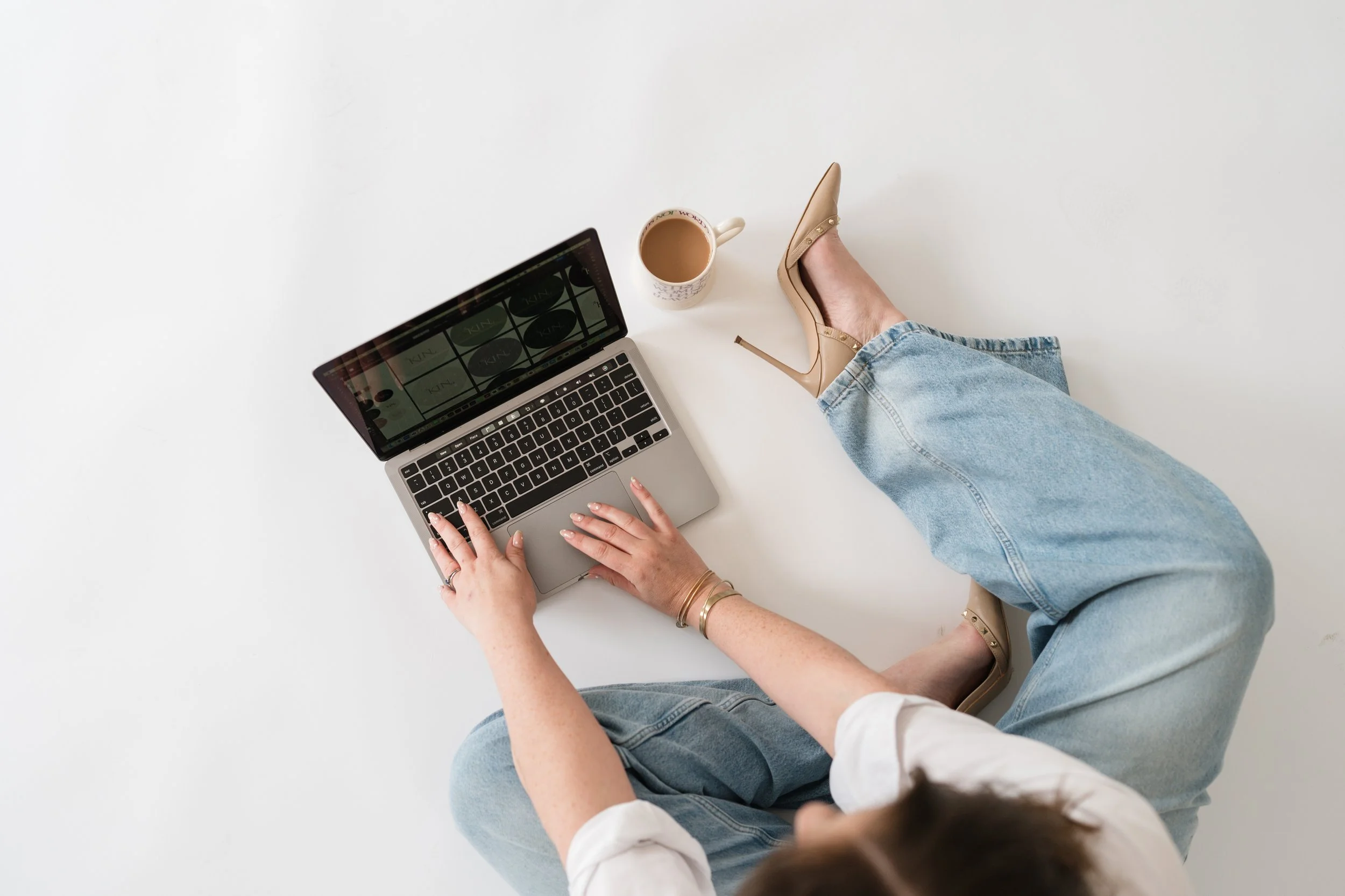 A person sitting on the floor with legs crossed, using a laptop, with a cup of coffee nearby, wearing beige high heels and light blue jeans.