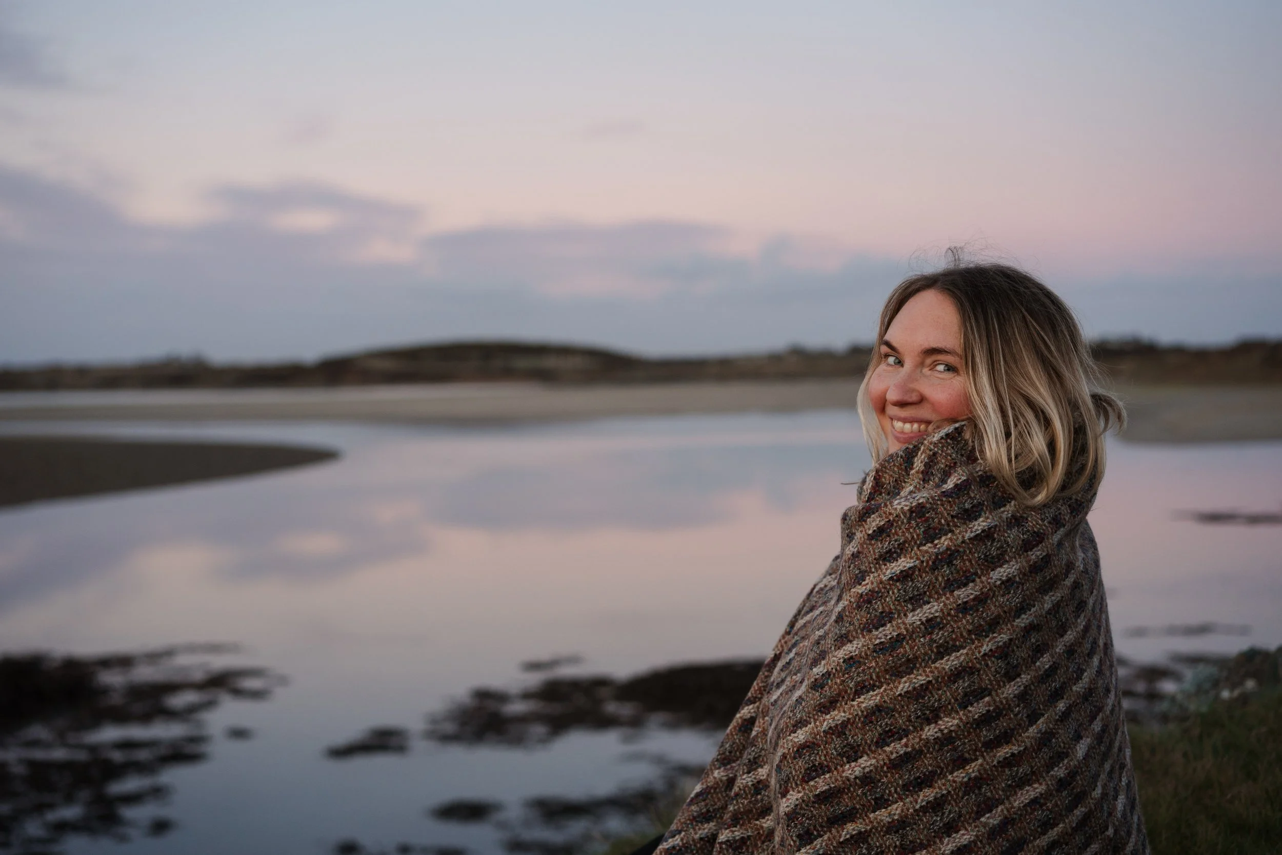 A young woman smiling while wrapped in a patterned blanket near a calm body of water at dusk, with hills and a cloudy sky in the background.