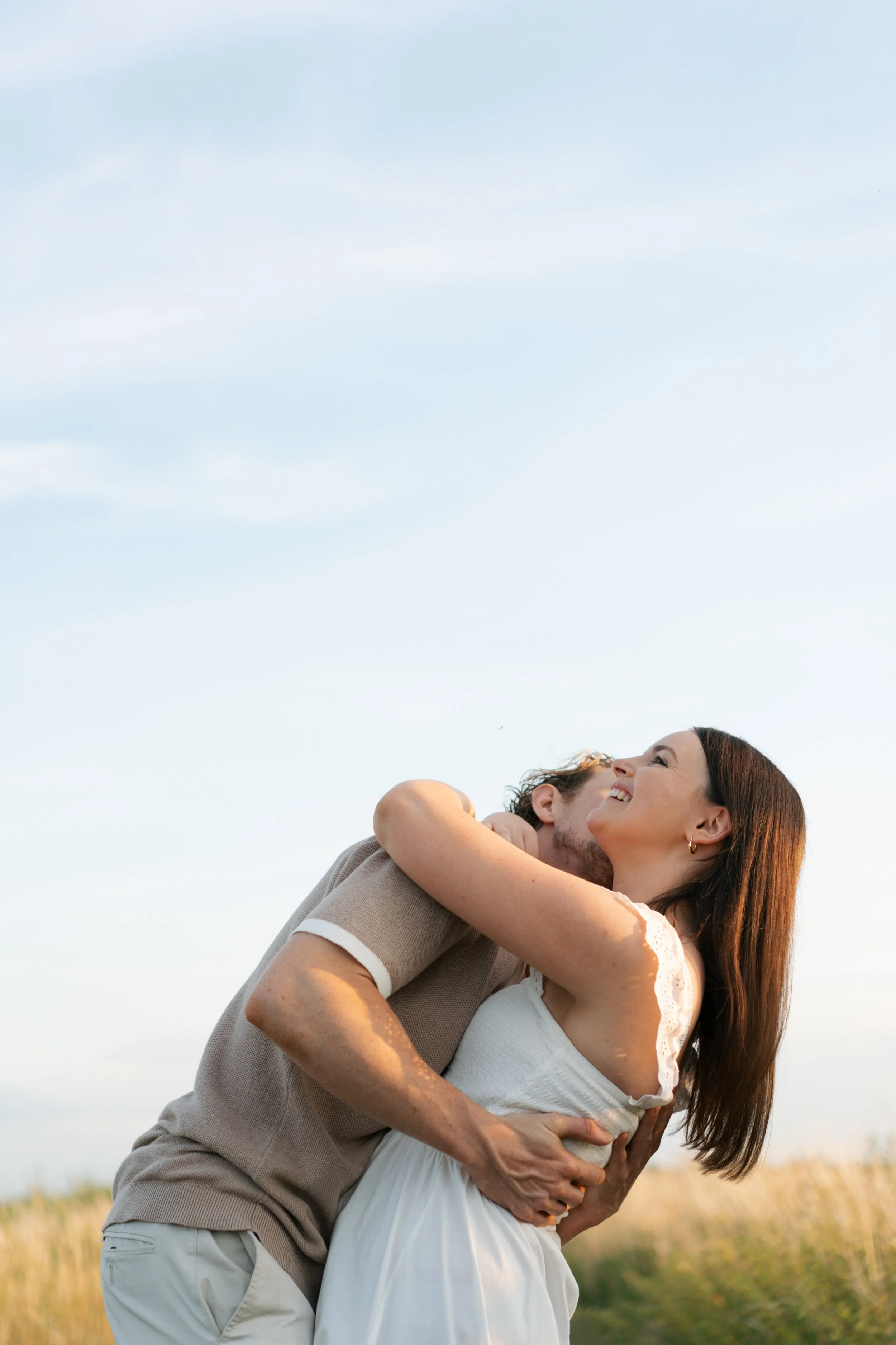 A couple hugging outdoors, with the woman smiling and looking up at the sky, in a field under a cloudy sky.