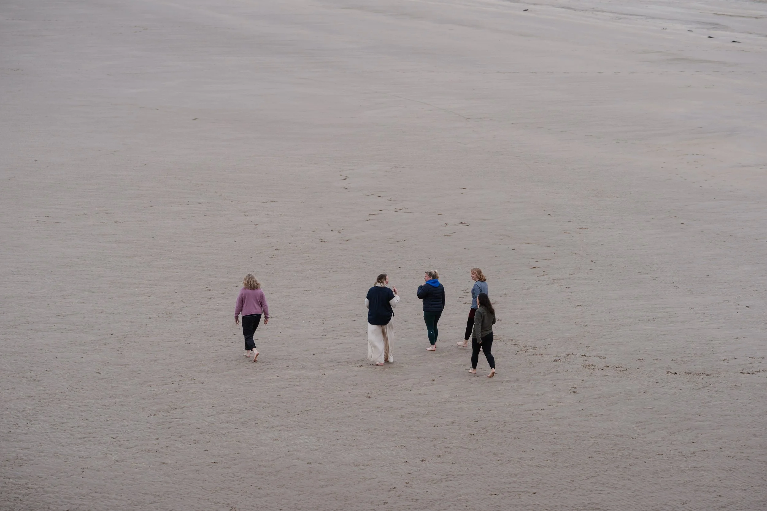Five women walking on a vast, sandy beach with no other features visible.