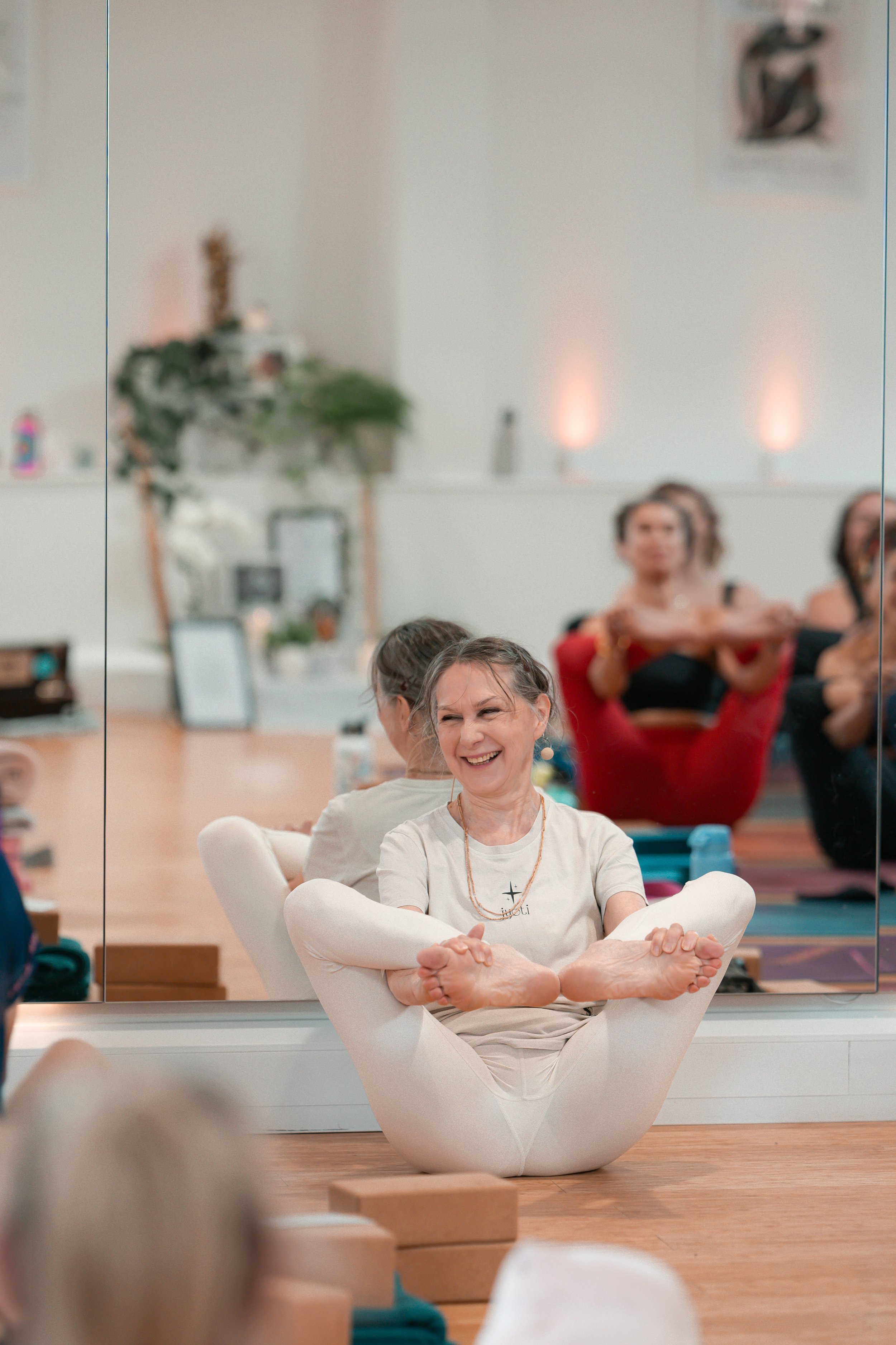 A woman sitting cross-legged on the floor, smiling joyfully during a yoga or meditation class, with reflective mirror in the background showing other participants in a similar pose.