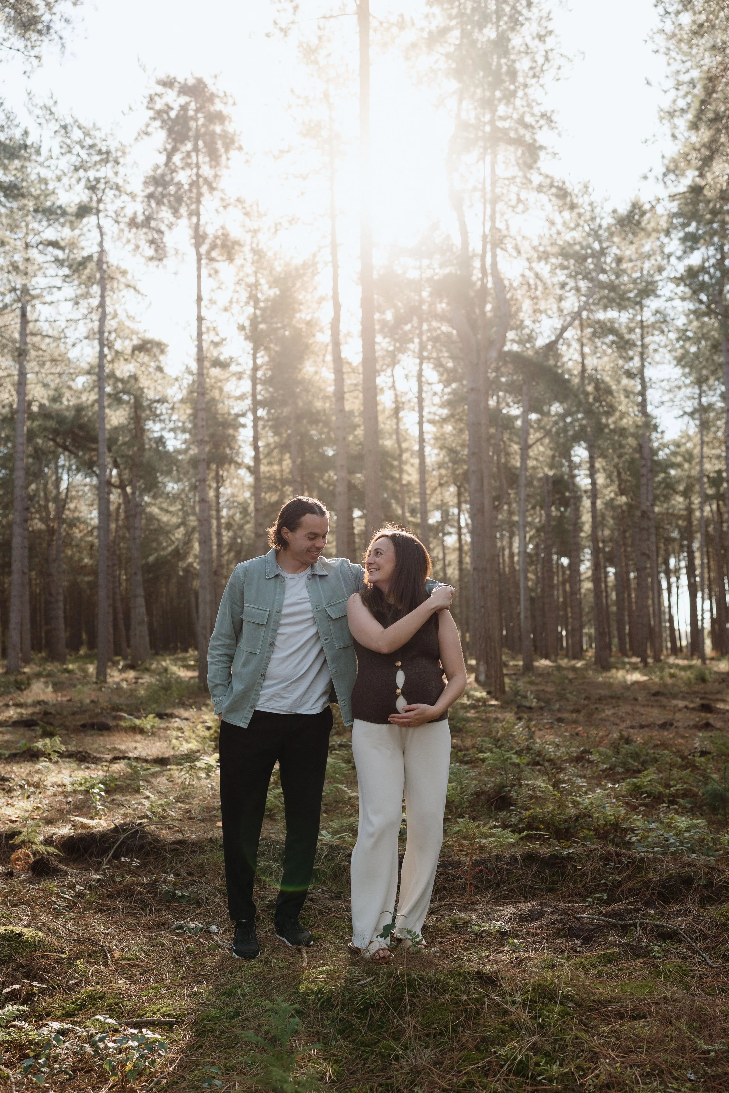 A couple walking in a forest, smiling at each other, with the woman holding her pregnant belly, during sunset.