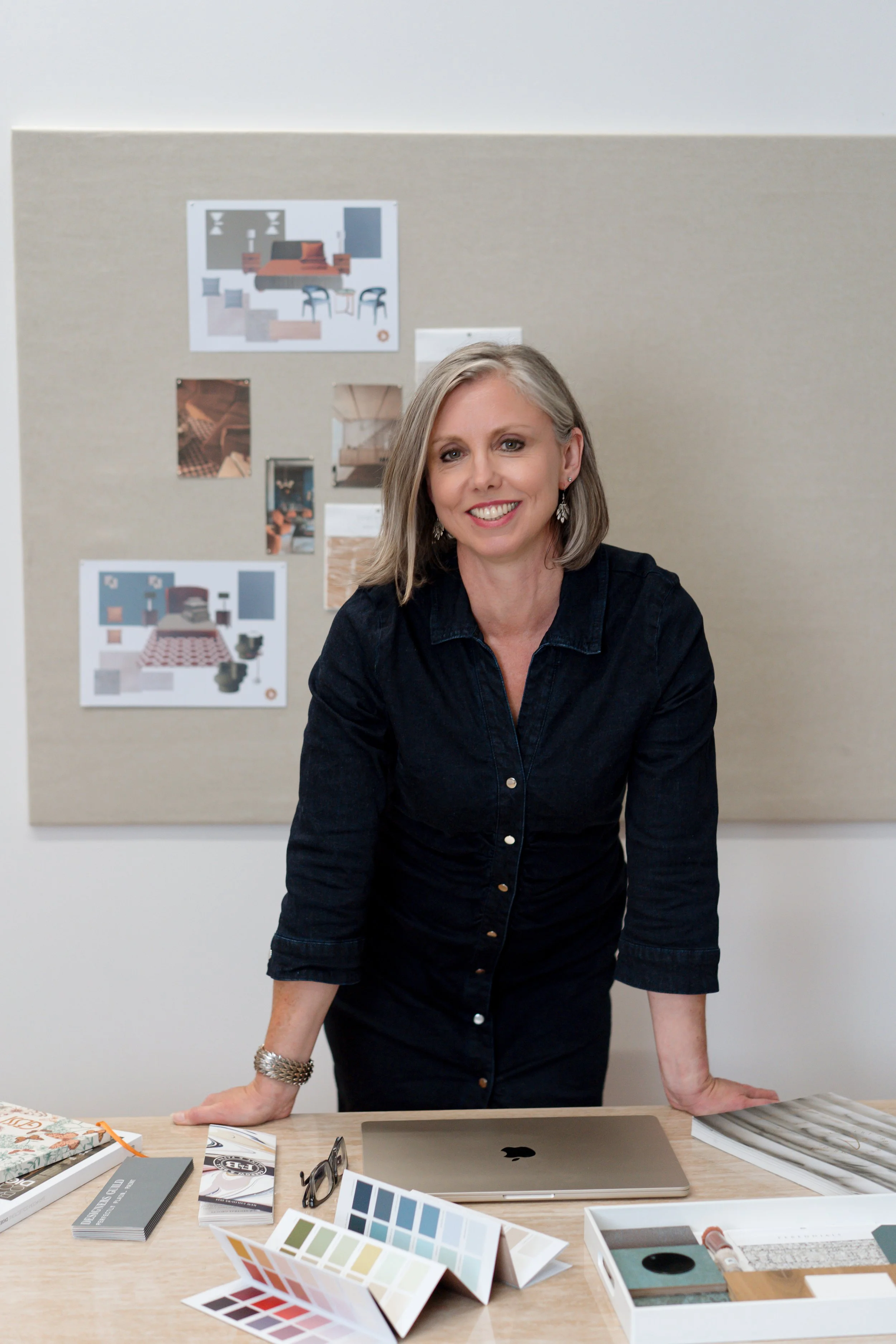 A woman with blonde hair wearing a black shirt, smiling and leaning on a desk in an interior design office. The desk has color swatches, a laptop, and design materials. A beige board with interior design sketches and photos is on the wall behind her.