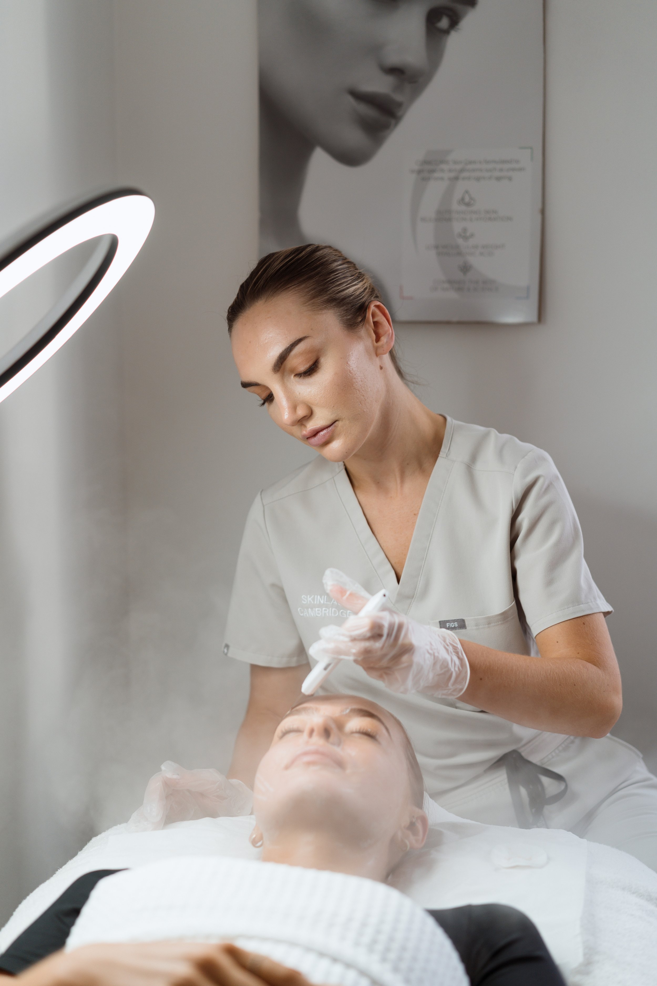 A woman receiving a facial treatment at a skincare clinic. The practitioner is using a device near the woman's face, both wearing gloves. The scene is well-lit with a ring light, and there is a large black-and-white portrait of a woman's face on the 