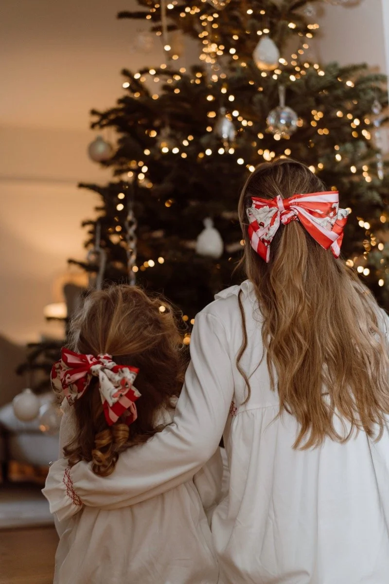 Two young girls with bows in their hair, sitting by a decorated Christmas tree with lights and ornaments, in a cozy indoor setting.