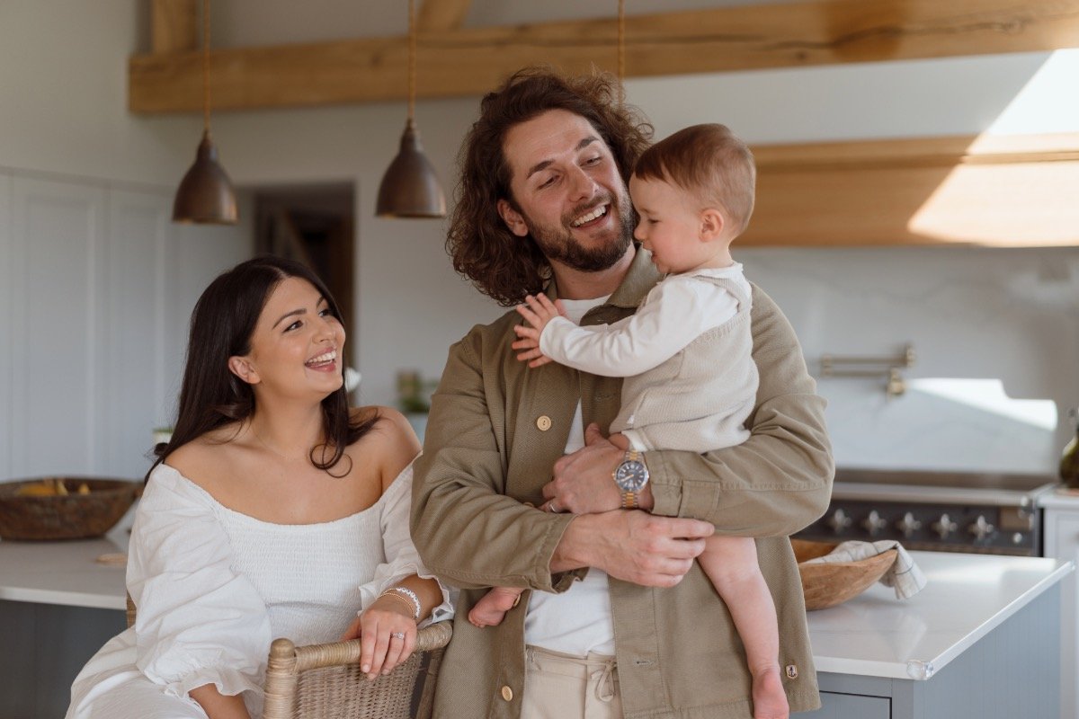 A family of three enjoying time together in a modern kitchen. The man with curly hair and beard is holding a smiling young boy, while a woman with long dark hair stands beside them, also smiling.
