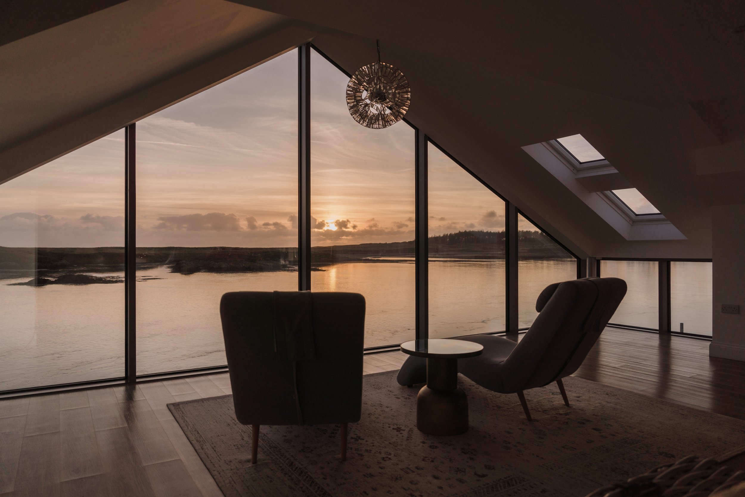 Interior of a modern room with large floor-to-ceiling windows showing a sunset over water and a distant shoreline, with a table and chairs inside.
