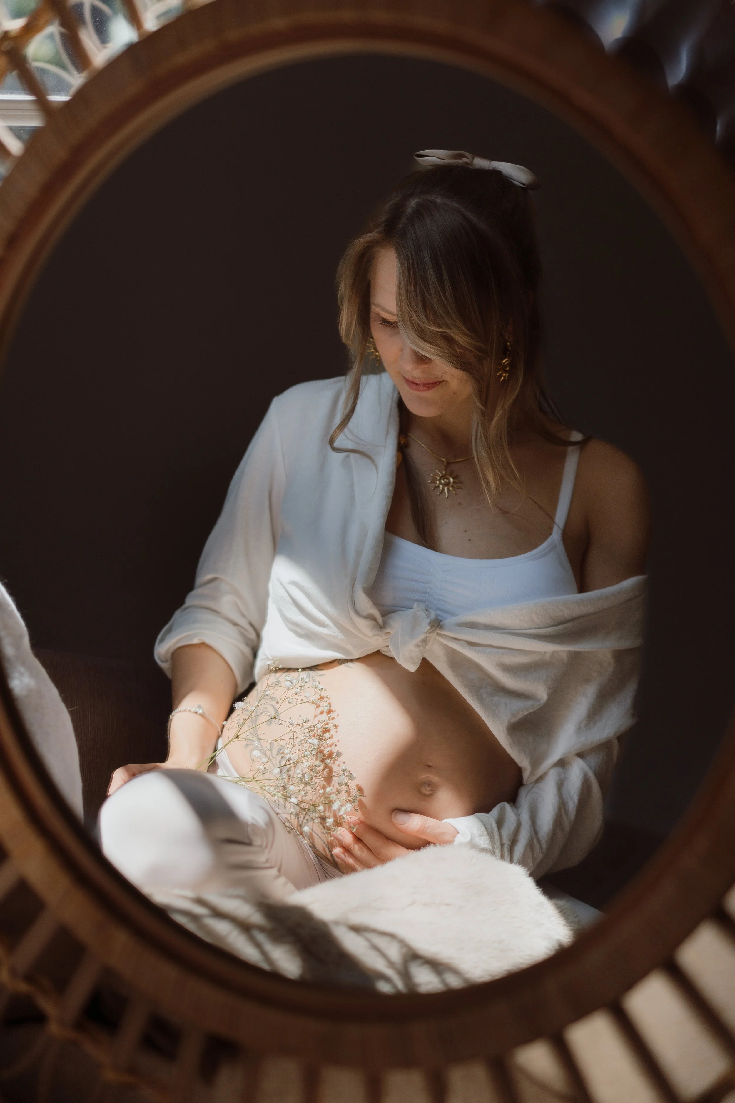 A woman looking down at her pregnant belly, which she gently touches, as seen through a round mirror. She is smiling and wearing jewelry and a white top, with flowers resting on her stomach.