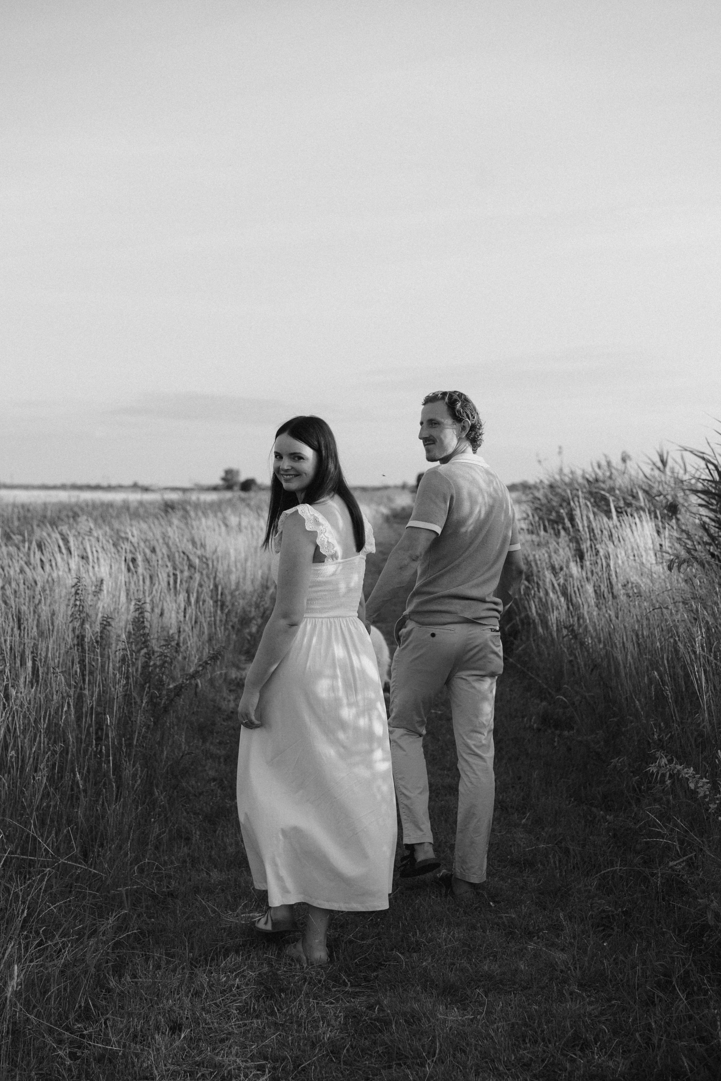 A black and white photo of a couple walking down a grassy path through a field of tall grasses, smiling and looking back over their shoulders.
