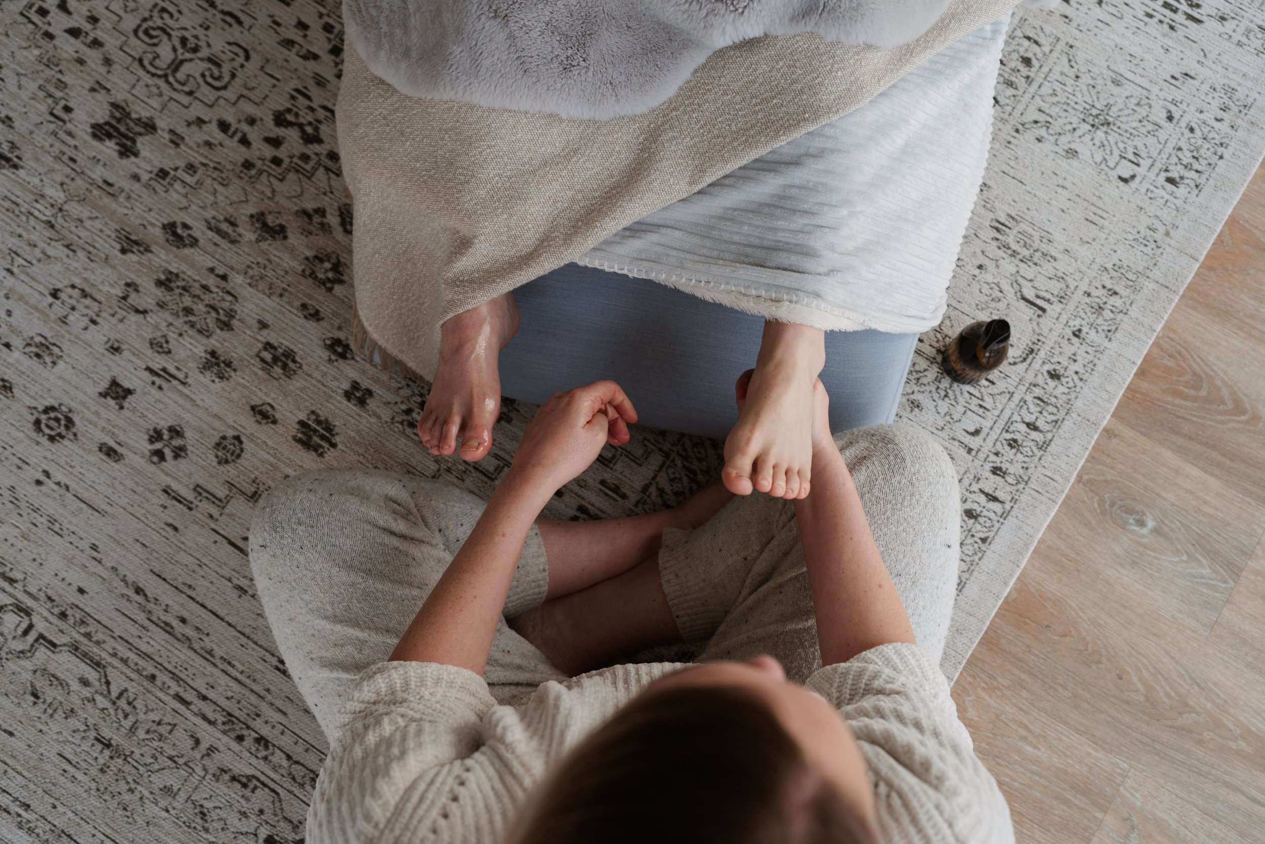 A person sitting on the floor with their legs crossed, holding another person's foot, while another person sits on a chair, engaged in a foot massage or tickling. There are rugs and a shoe on the floor nearby.