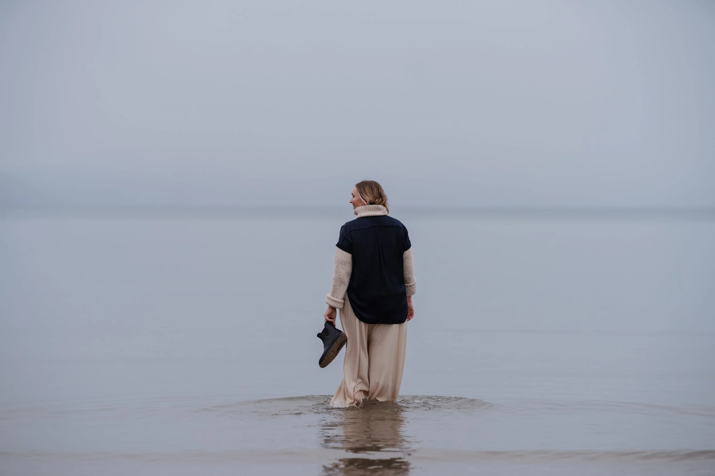 Woman walking in shallow water at the beach, holding a pair of black shoes, with calm ocean and overcast sky in the background.
