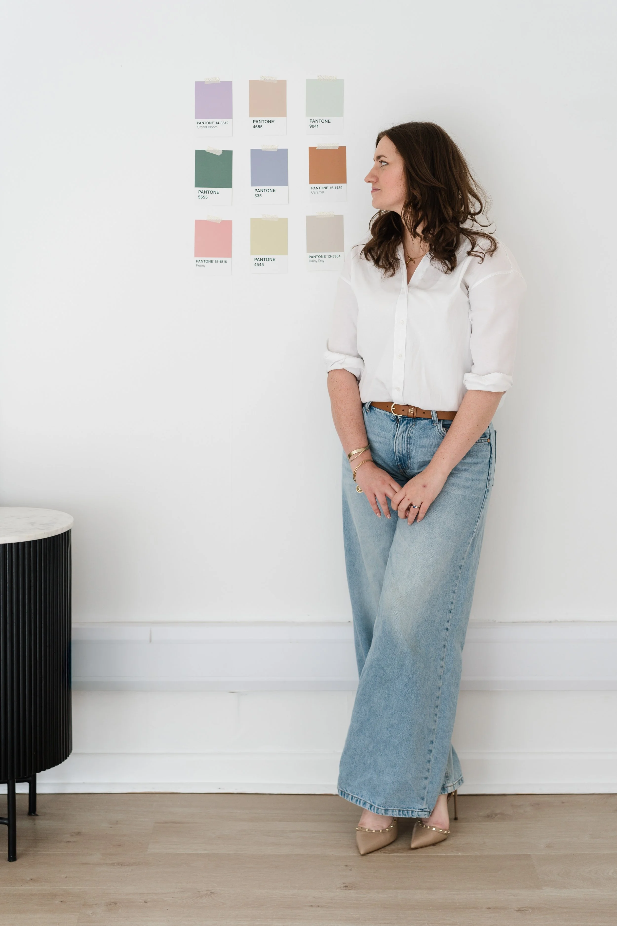 A woman with brown hair wearing a white blouse and wide jeans standing in a minimalist white room looking to the right, with color swatches on the wall behind her.