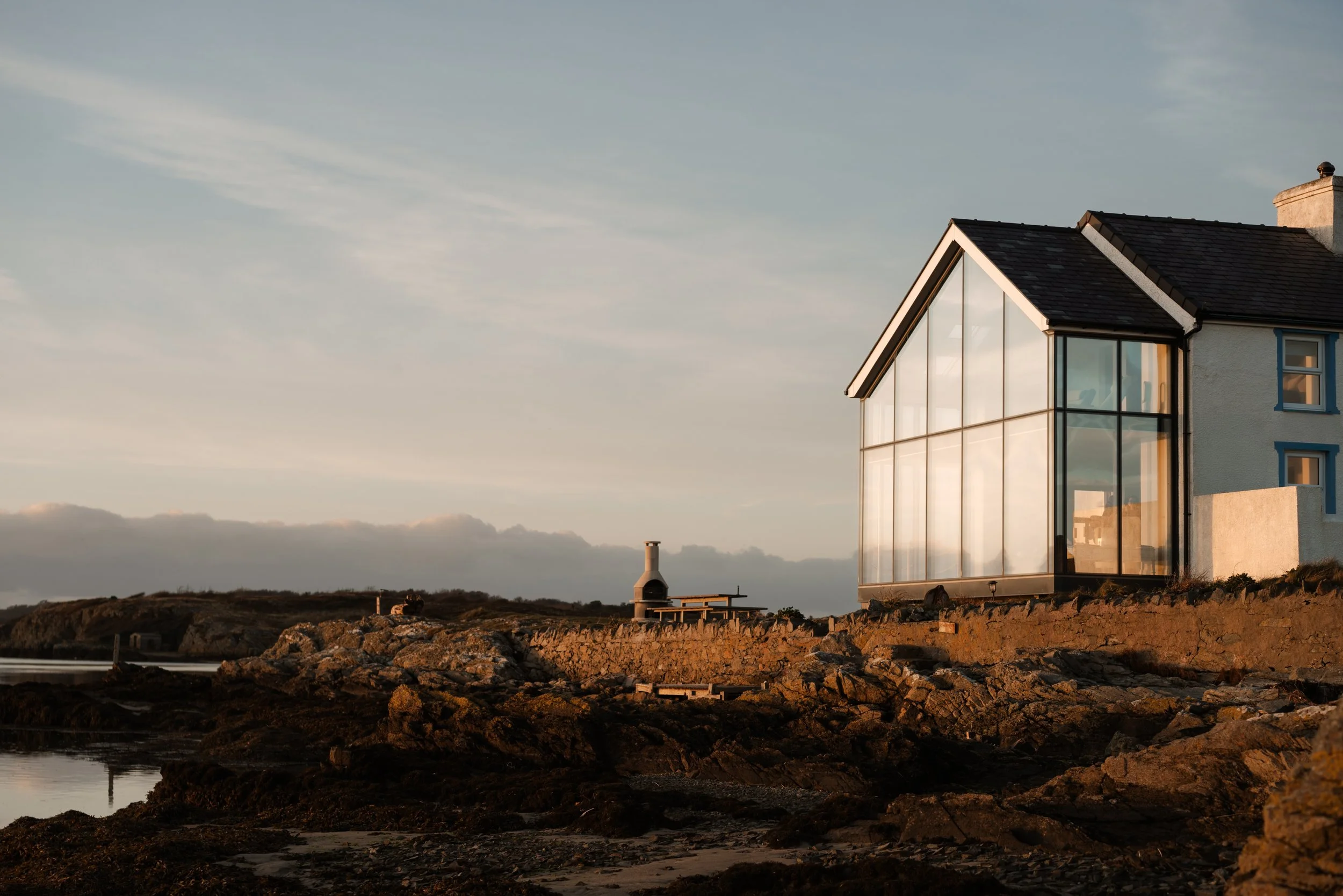 A house with large glass windows on a rocky shoreline during sunset