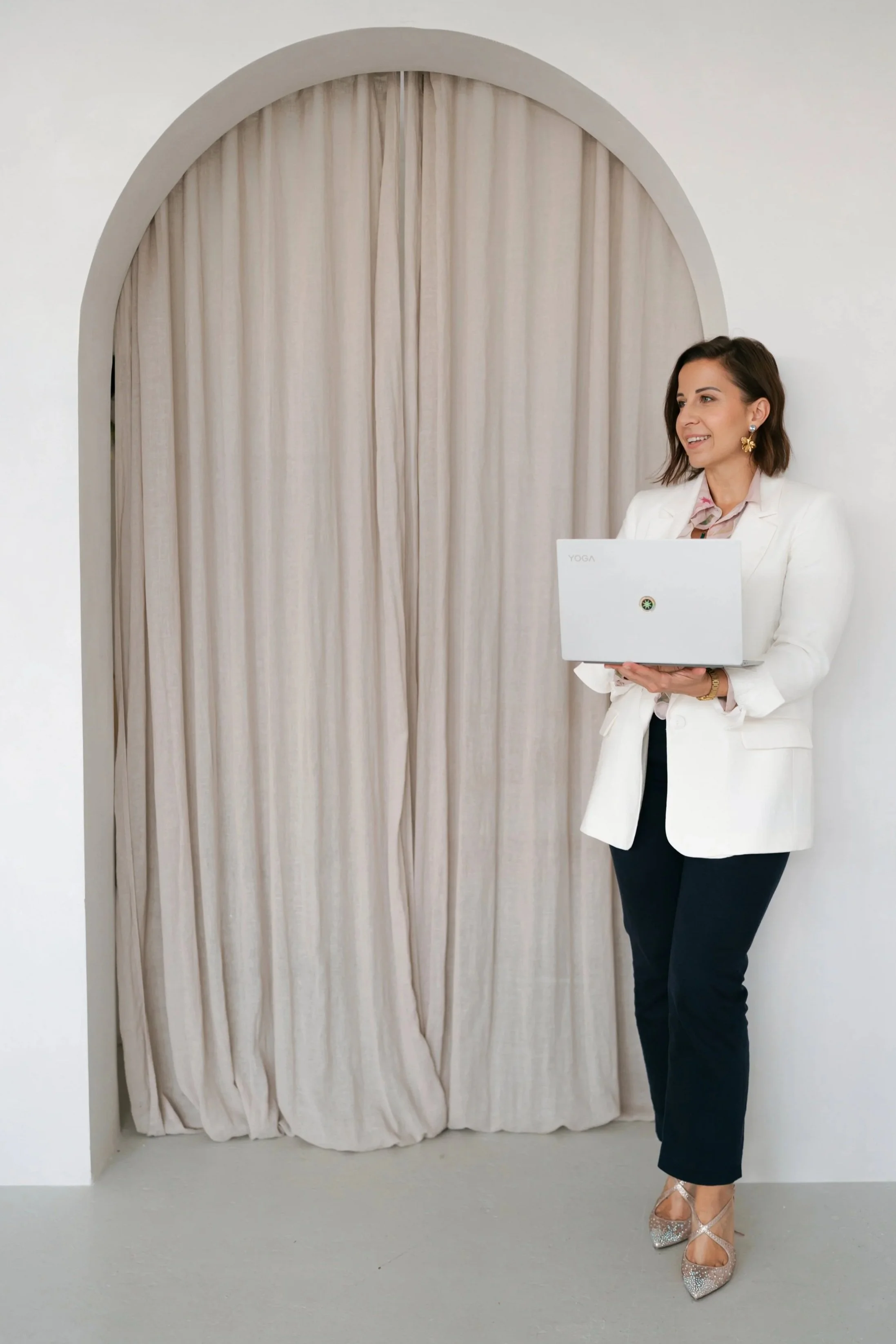 A woman in a white blazer and dark pants holding a laptop in front of beige curtains inside a room with white walls.