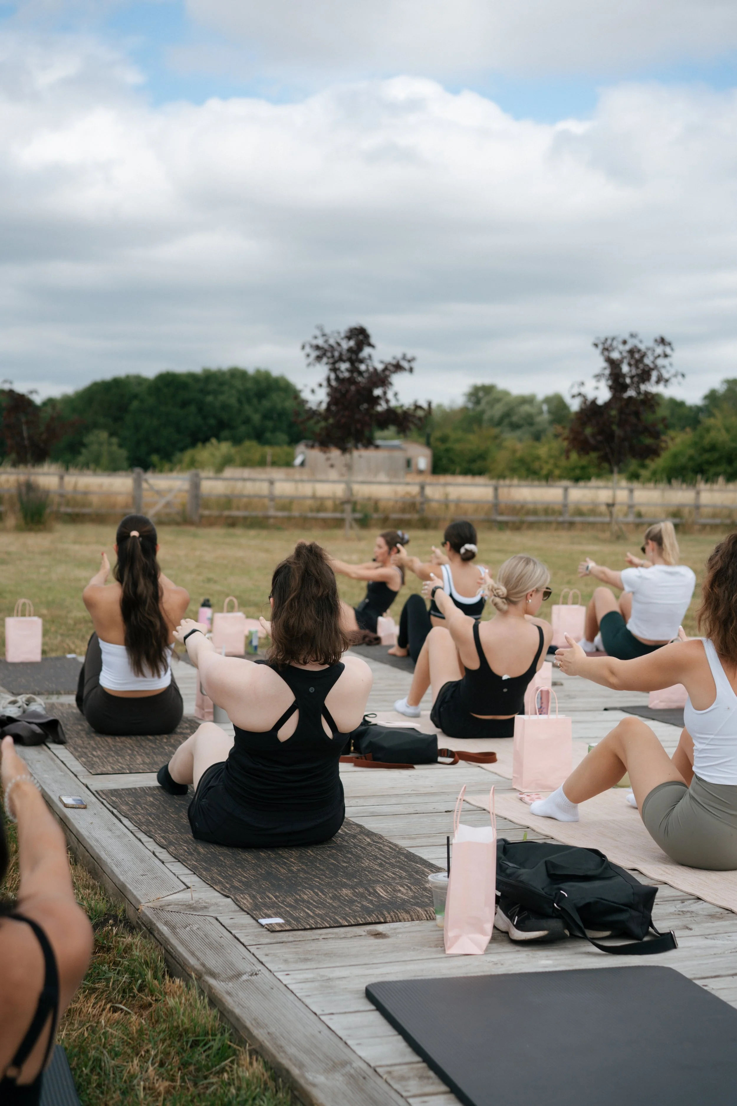 People participating in an outdoor yoga class on a wooden platform in a field, with trees and cloudy sky in the background.