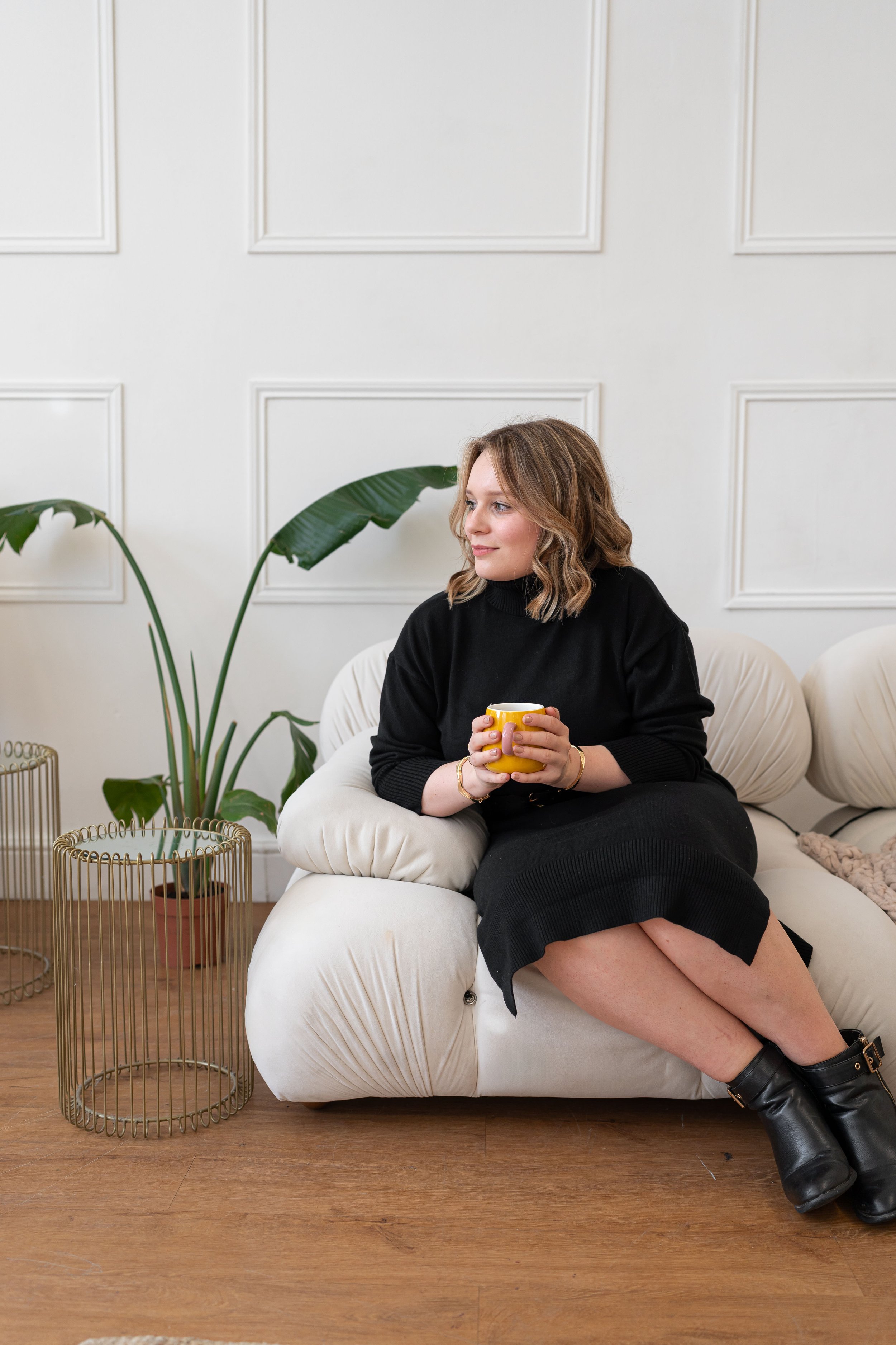 A woman with shoulder-length wavy hair sitting on a white sofa, holding a yellow mug, in a room with white walls, green plants, and wooden flooring, wearing a black sweater, black skirt, and black boots.