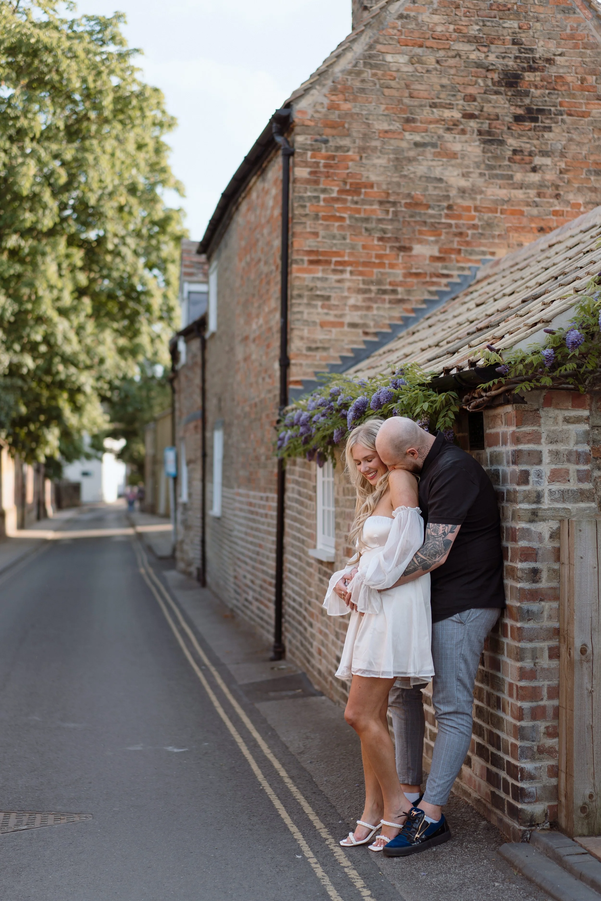 A couple standing close together on the sidewalk against a brick wall, smiling and embracing on a sunny day.