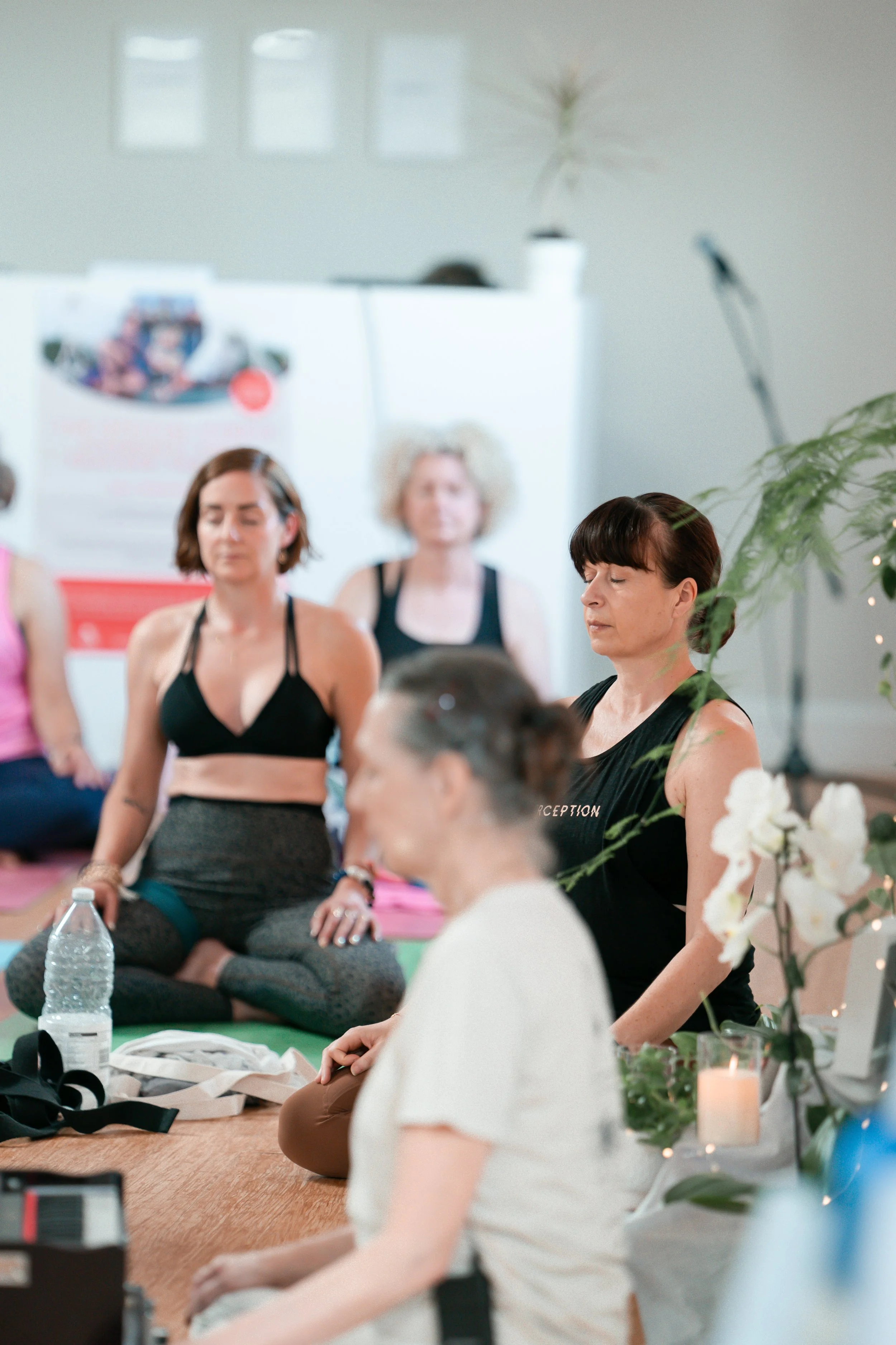 Group of women sitting cross-legged on the floor in a meditation pose with their eyes closed during a meditation session in a bright room with plants and candles.