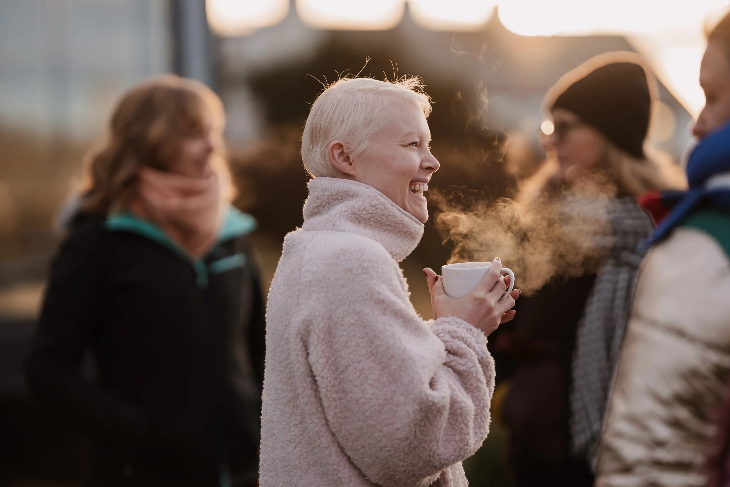 A woman with short blonde hair smiling and holding a steaming cup, surrounded by other people outdoors during sunset in warm clothing.