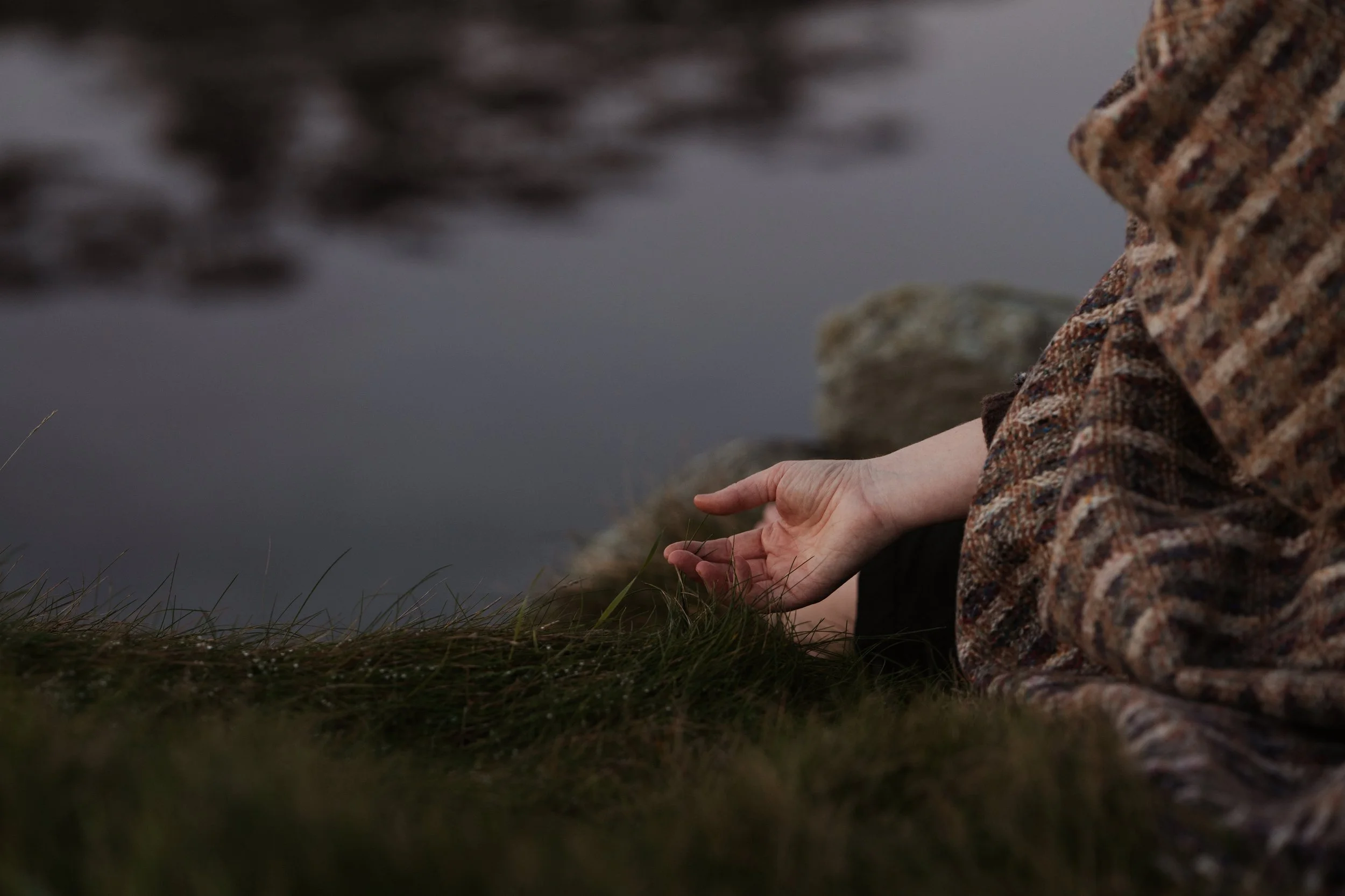 A person sitting by a pond or lake, wearing a patterned sweater, with a hand resting on the grass near the water, rocks in the background.