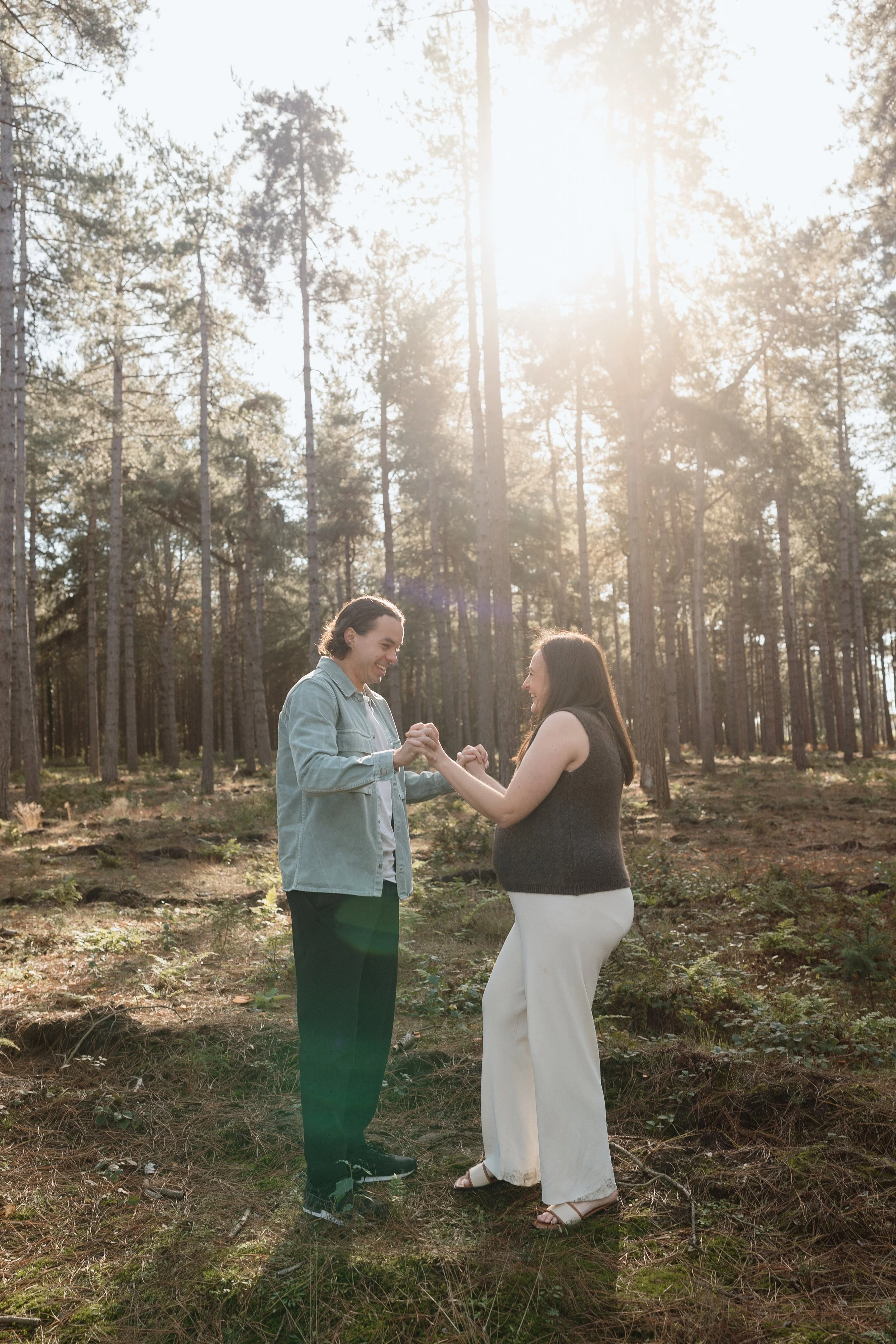A couple holding hands and smiling at each other in a sunlit forest.