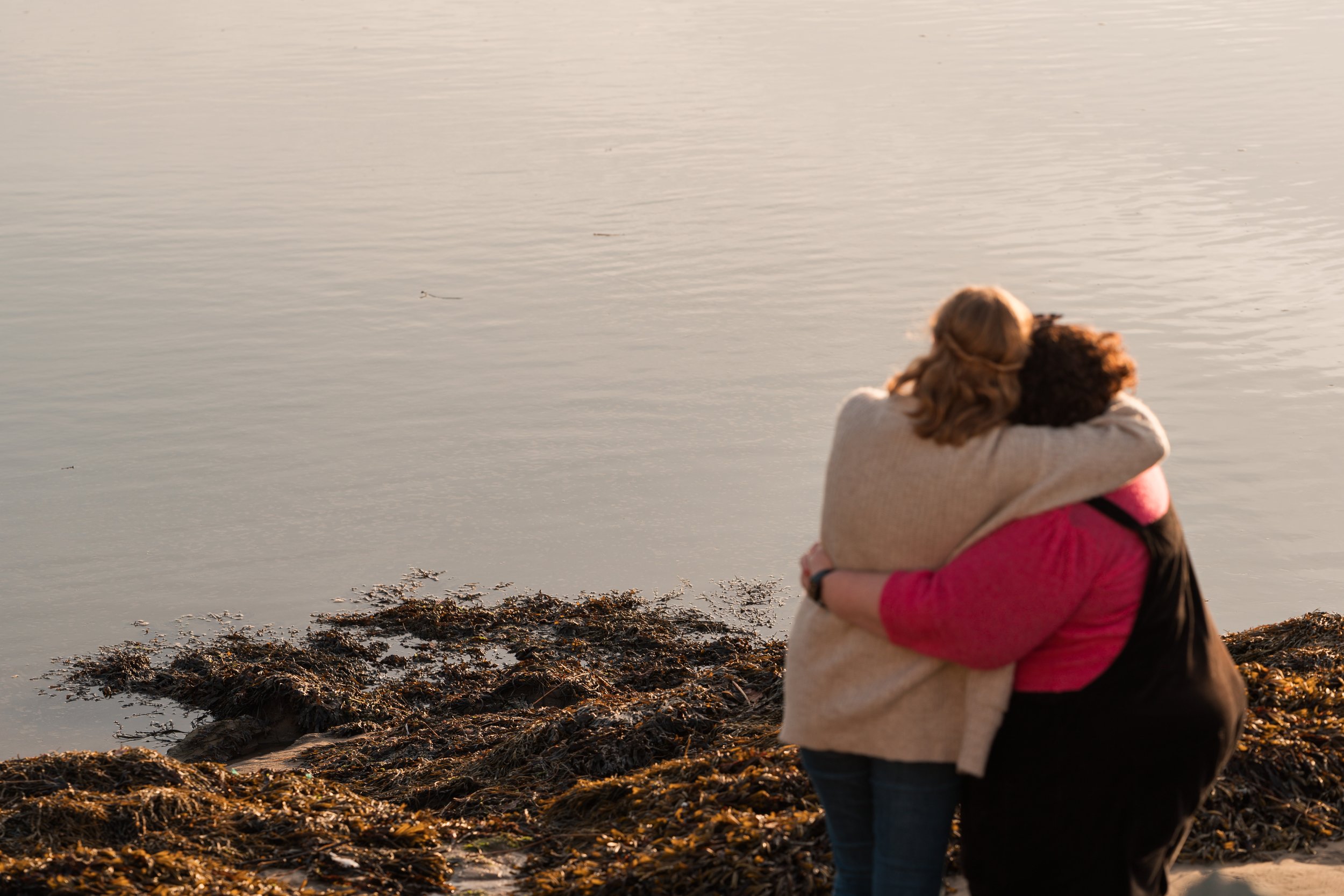 Two women embracing on a beach near the water during sunset or sunrise.
