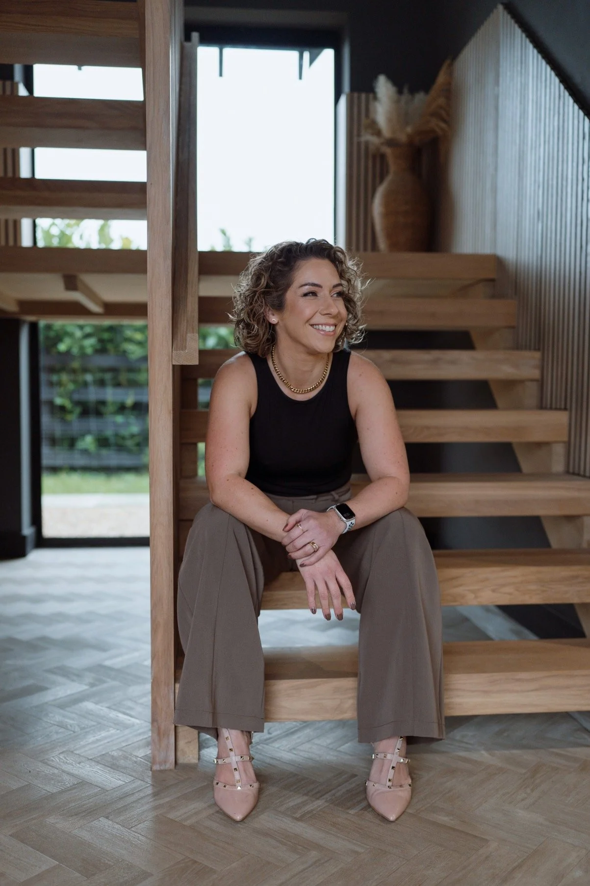 A smiling woman with curly hair, wearing a black sleeveless top, beige wide-leg pants, pink shoes, and a smartwatch, sitting on wooden stairs inside a modern home.