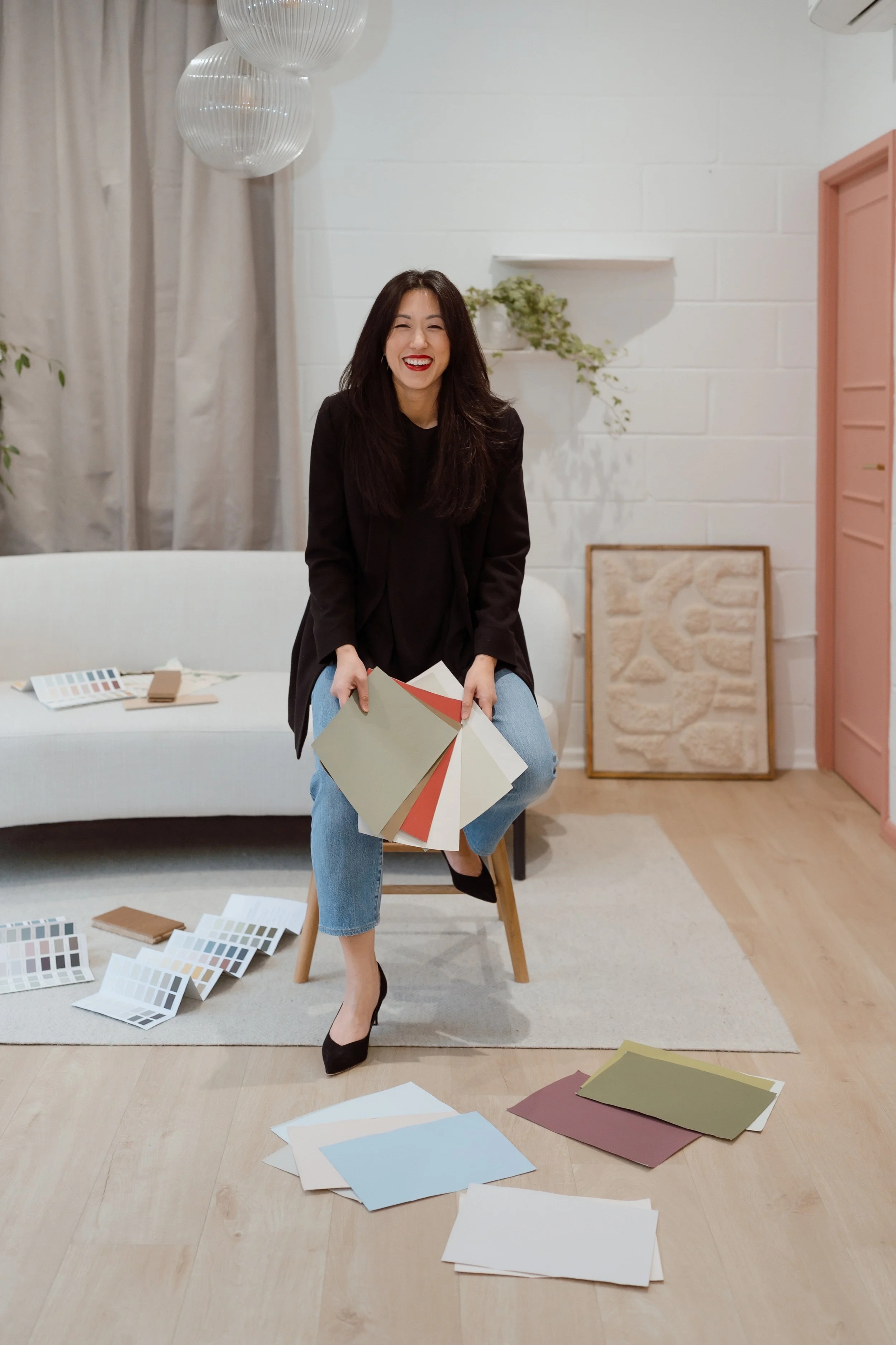 A woman sitting on a chair holding paint sample cards, with more color swatches and sample sheets on the floor and sofa in a modern, decorated room.