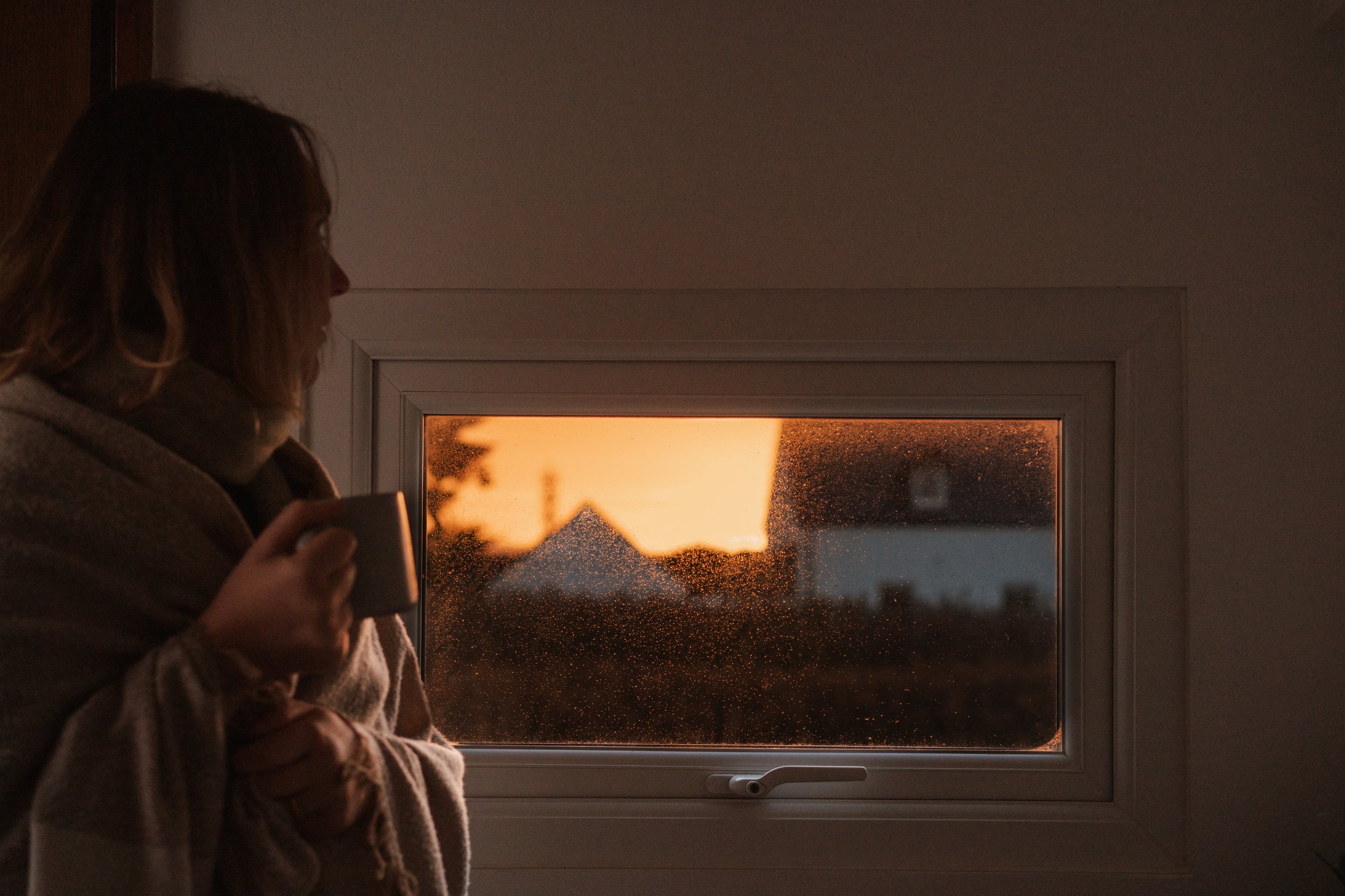 A person with shoulder-length hair, wrapped in a blanket, holding a mug, looking out a window at sunset with a silhouette of trees and buildings.