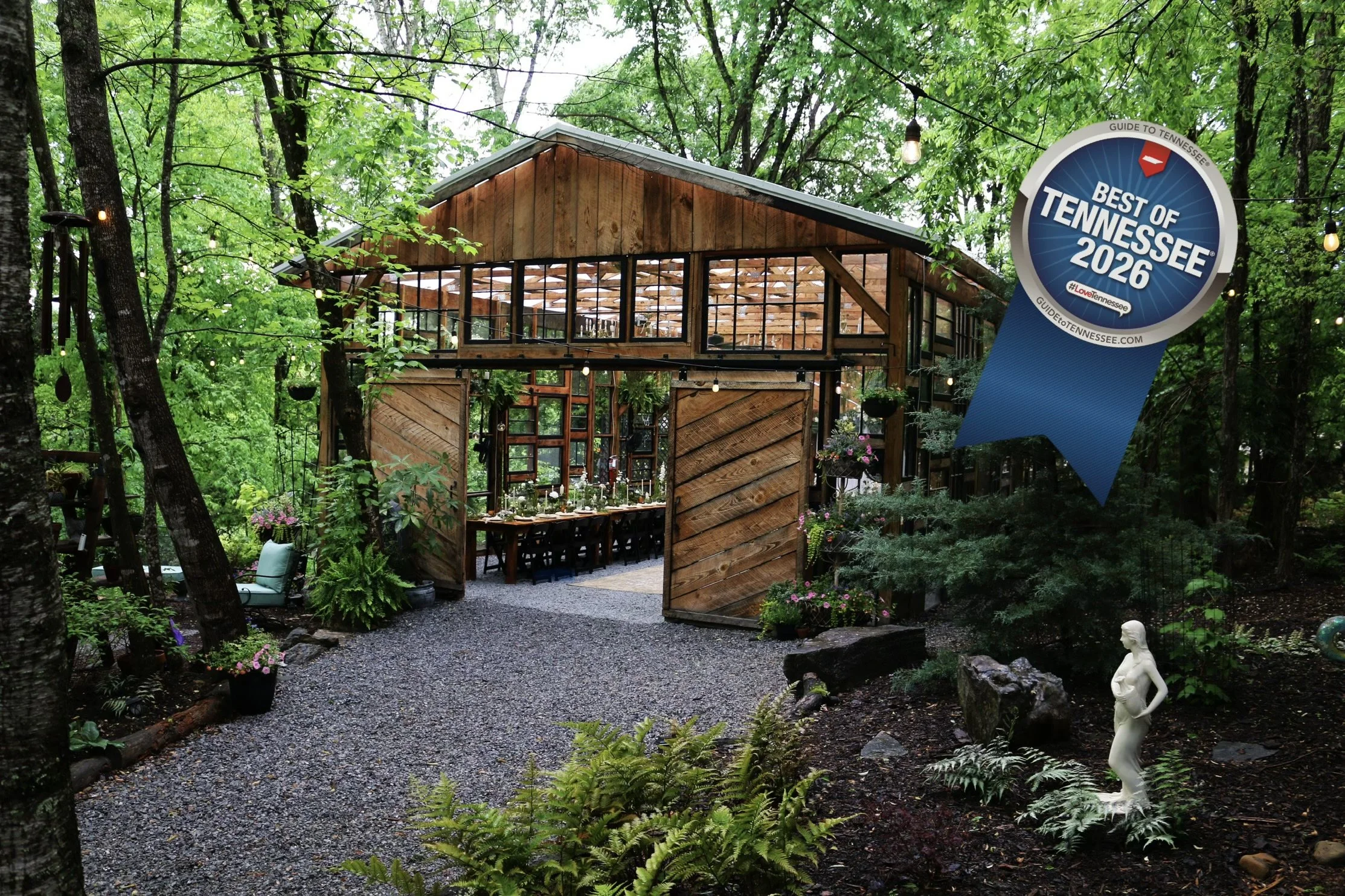 Wood-and-glass rustic building surrounded by lush forest, gardens, string lights, and a large Best of Tennessee 2026 sign.