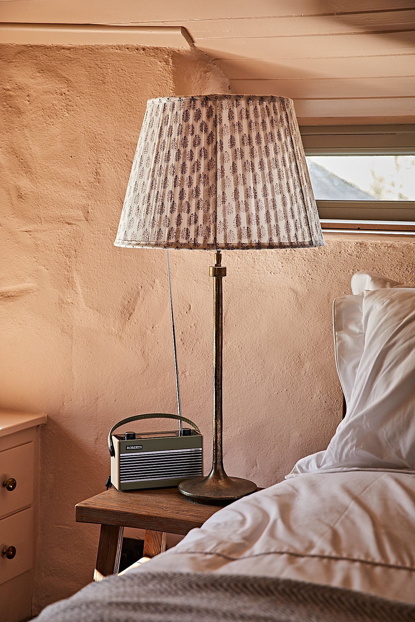 A bedroom scene in luxury holiday let The Dorset Nook with a wooden bedside table holding a vintage-style table lamp with a patterned lampshade and a small portable radio. 