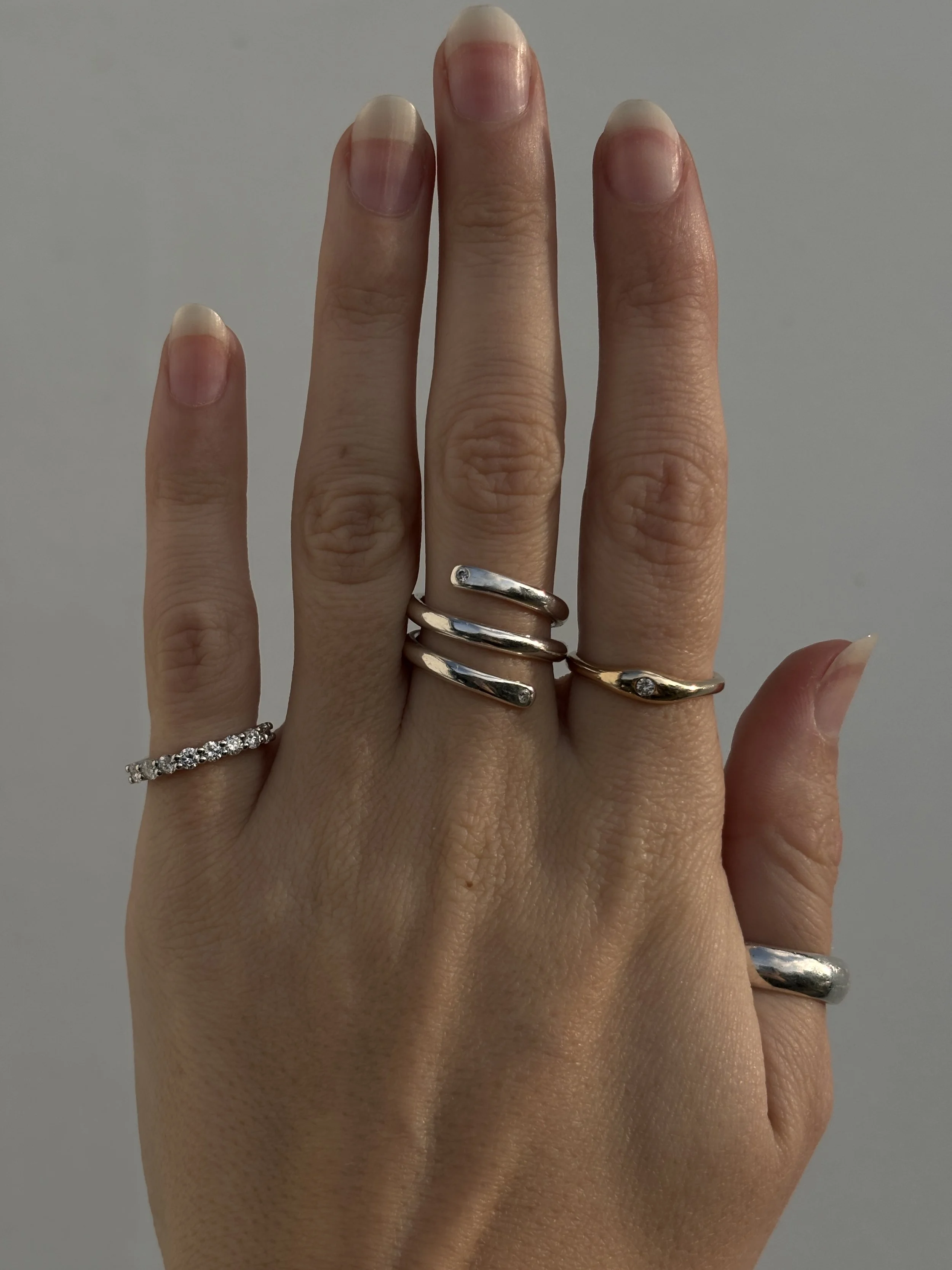 A person's hand with multiple rings, including a diamond band, a triple-band ring, and a gold ring with a small gemstone, displayed against a plain background.