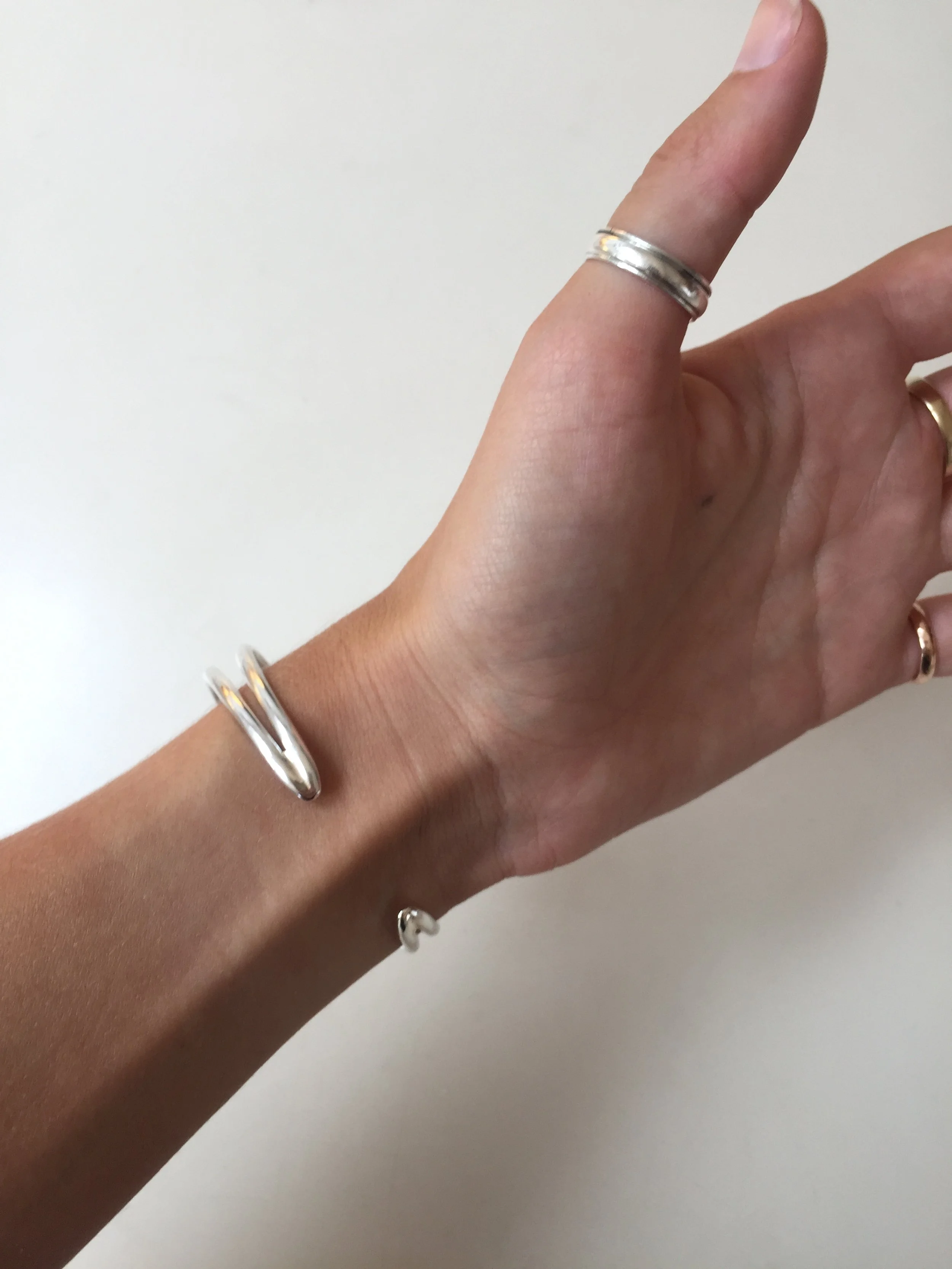A hand wearing multiple metallic rings and a metallic bracelet, with the fingers slightly bent and positioned against a plain white background.