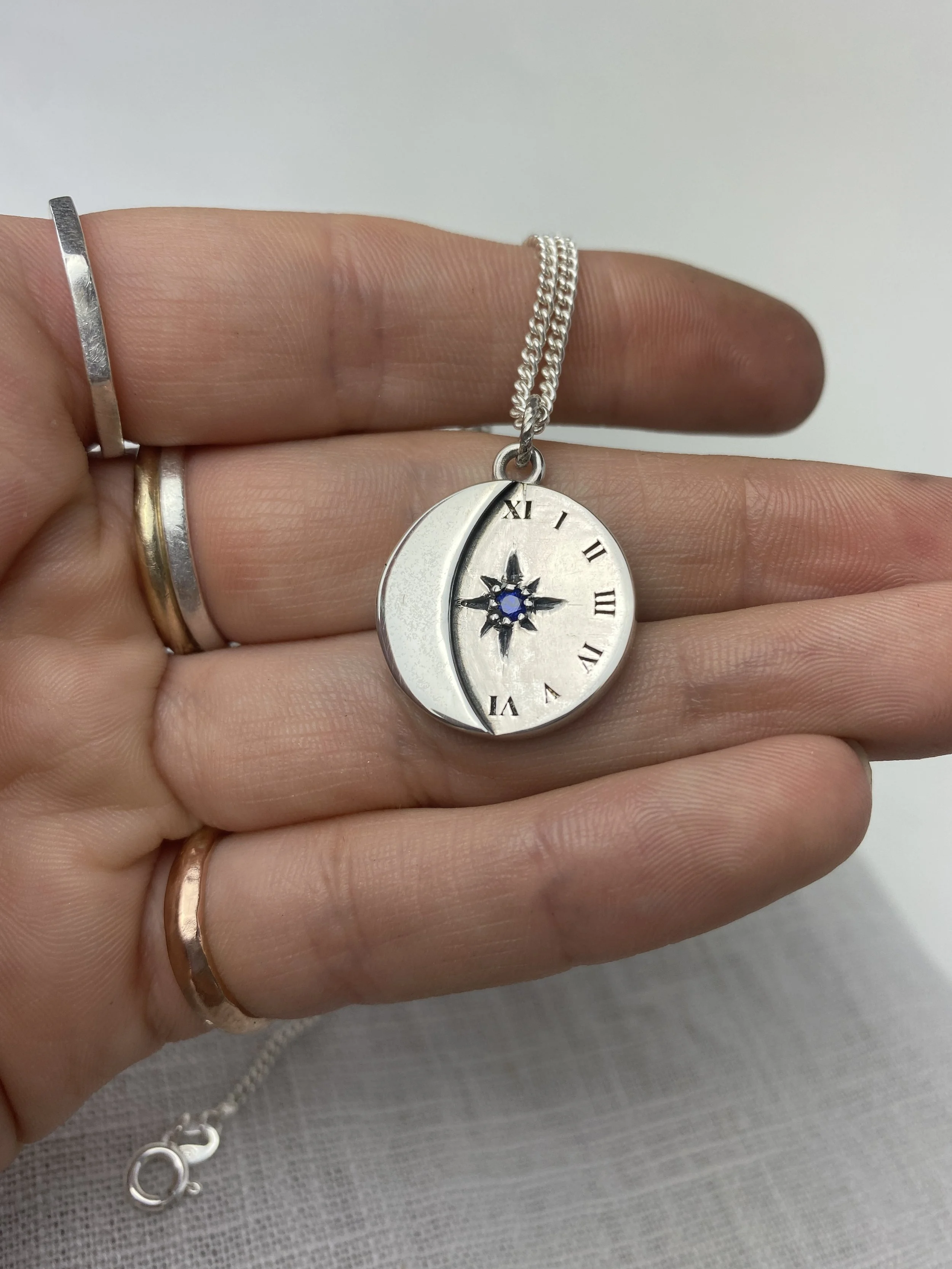 A hand holding a silver crescent moon and star-shaped pendant necklace, with Roman numerals inscribed on it, a blue gemstone in the center of the star, and multiple rings on the fingers.