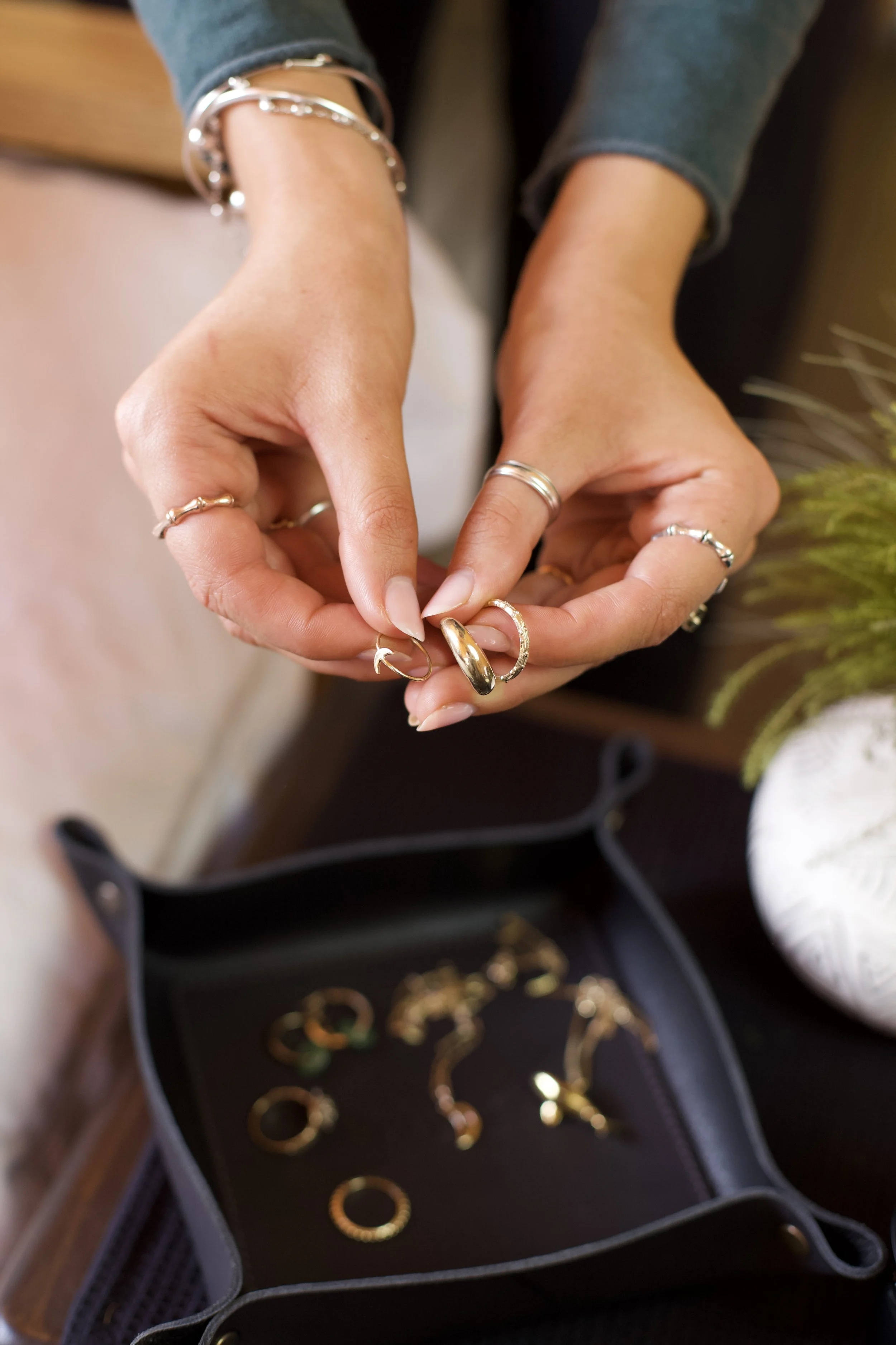 Persones handen met verschillende sieraden, zoals ringen en een armband, terwijl ze verschillende gouden en zilveren sieraden vasthouden, met een sieradenkistje met rings en hangers op de achtergrond.