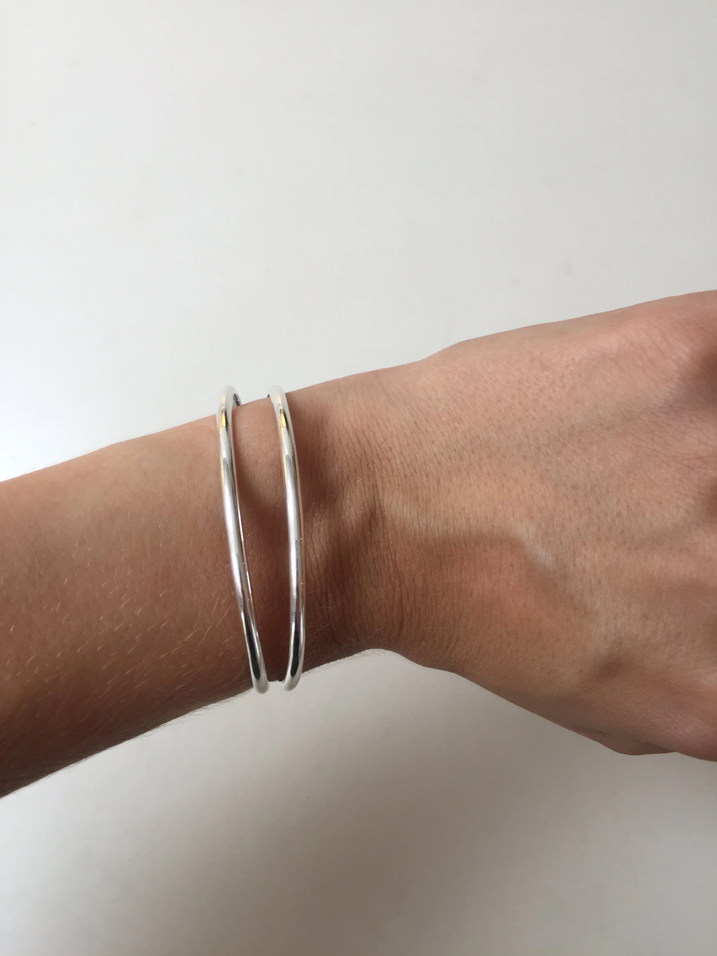 A person's wrist wearing two thin silver bangles, positioned against a plain white background.