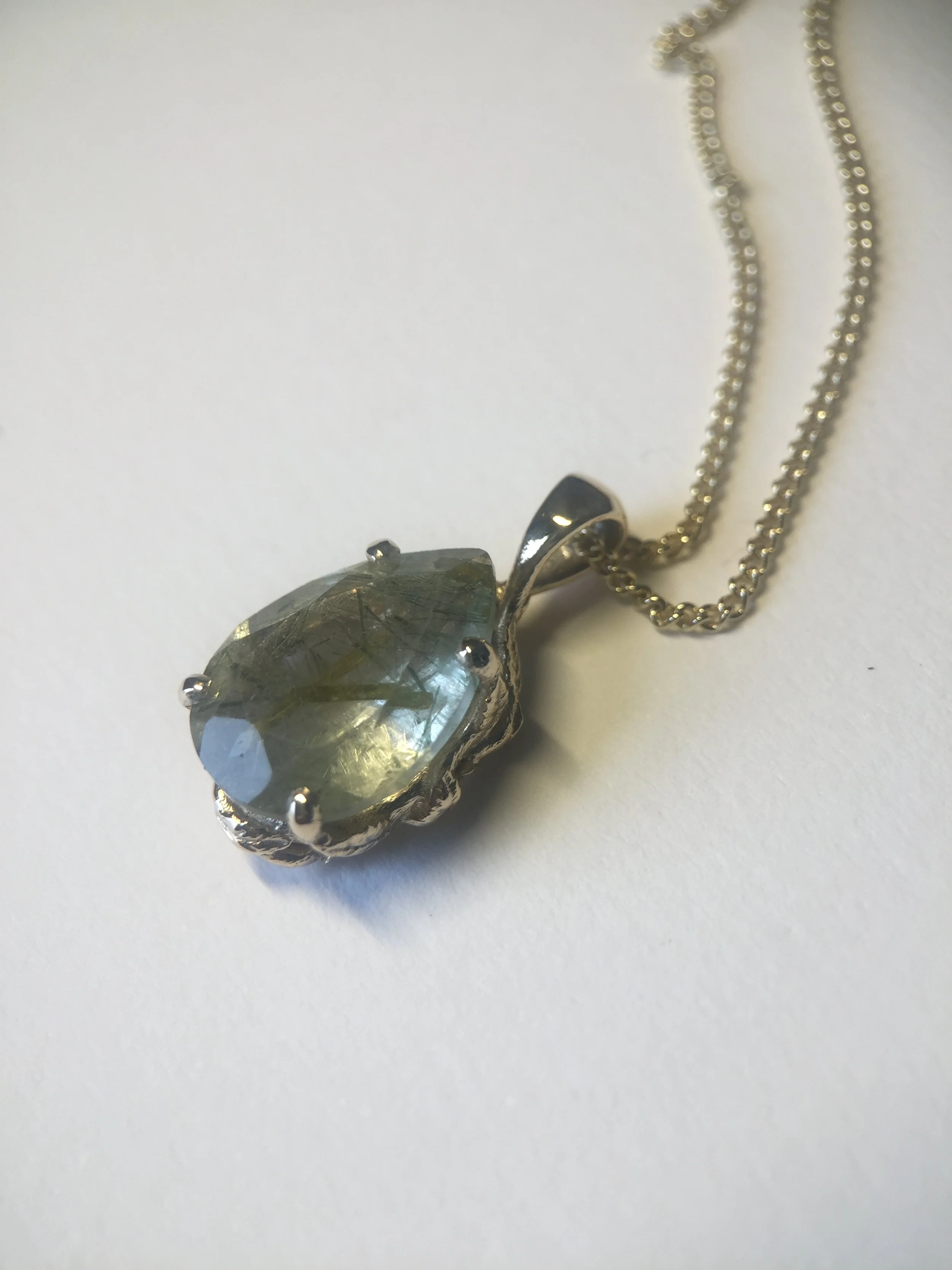 Pendant with a large, faceted, grayish-green gemstone in a silver setting, attached to a silver chain on a white background.