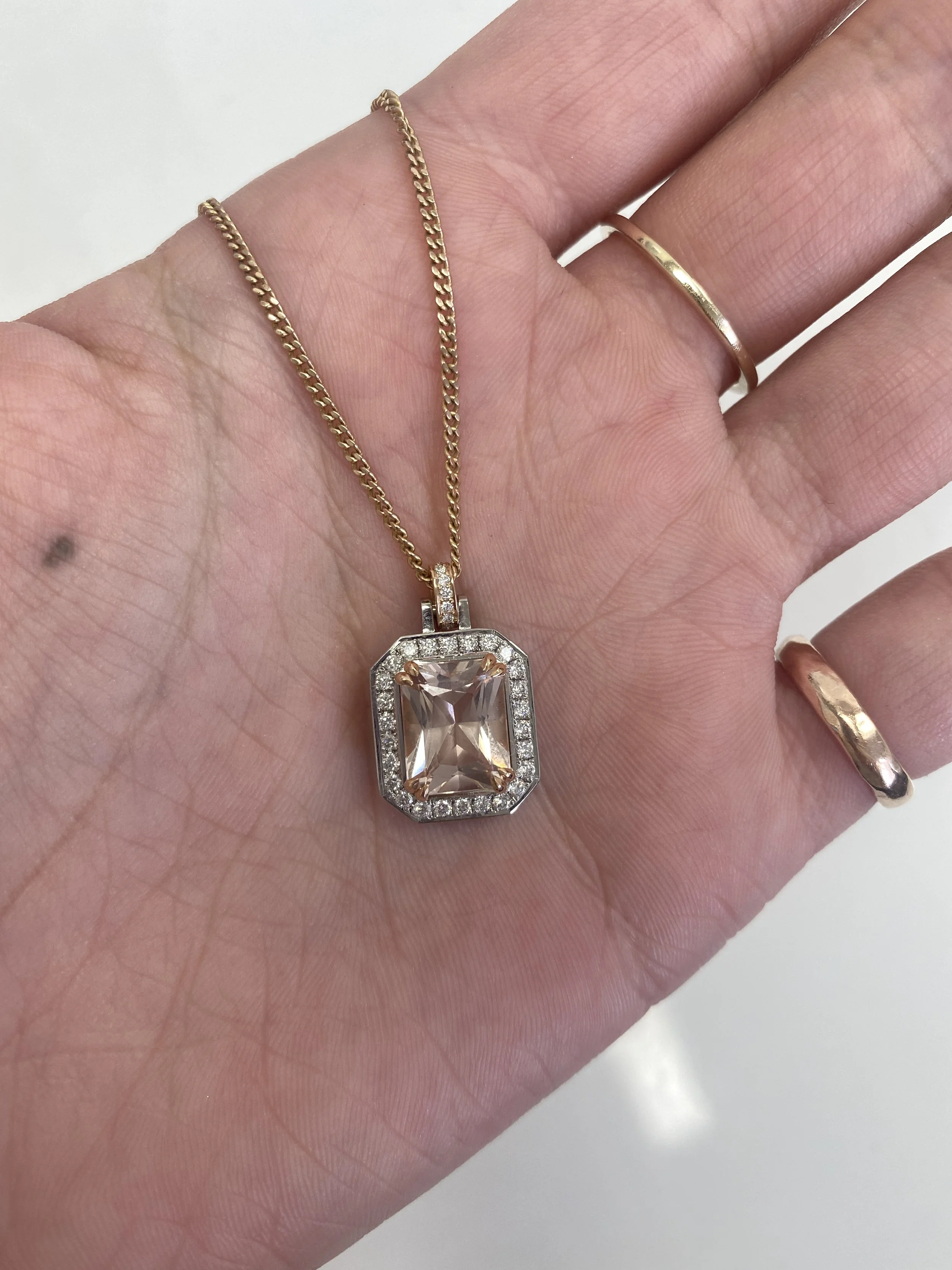 Gold necklace with a large rectangular gemstone pendant surrounded by small diamonds, held in a person's hand with rings.