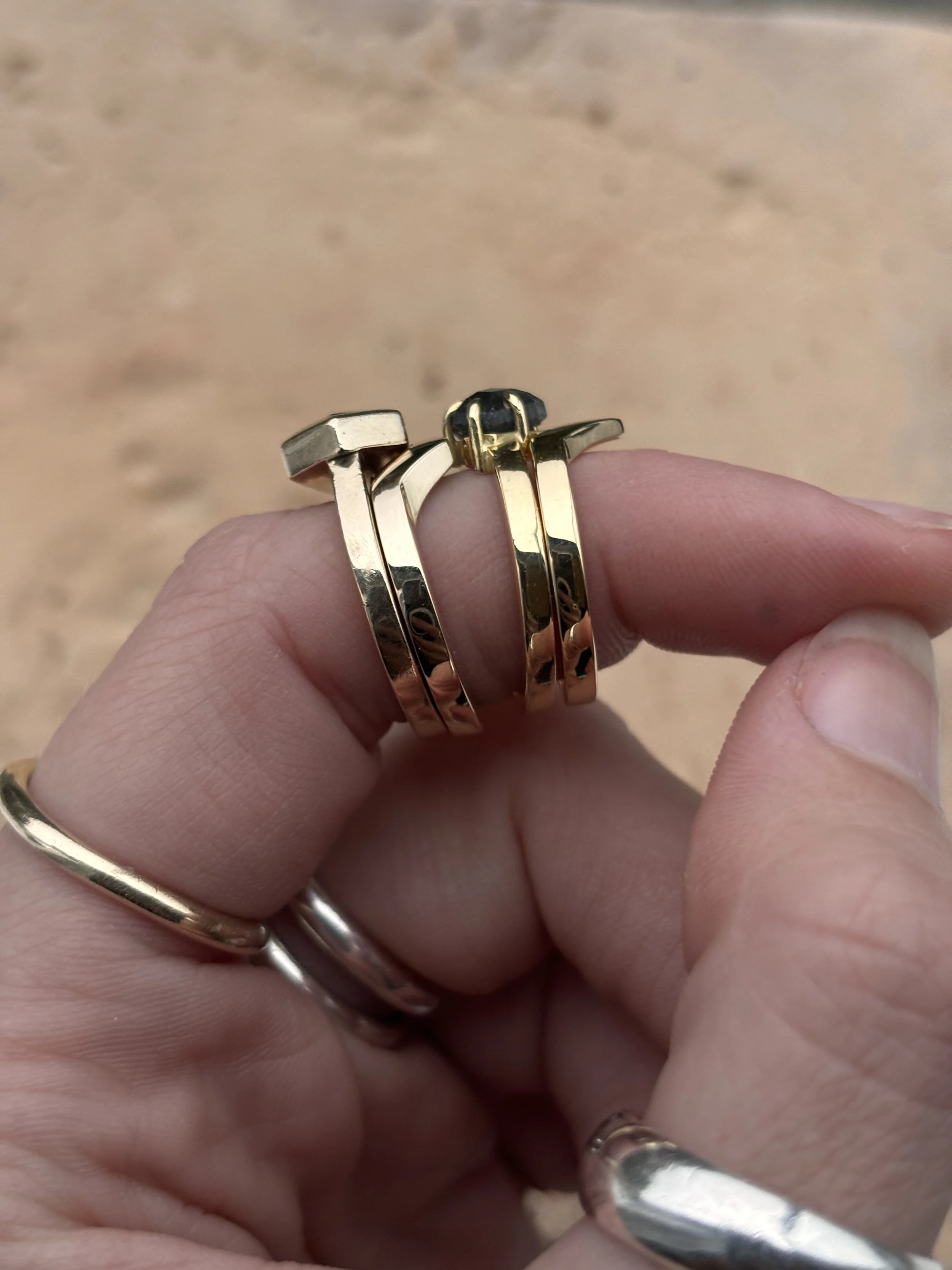 A person's hand holding a gold ring with multiple bands and a black stone on a simple background.